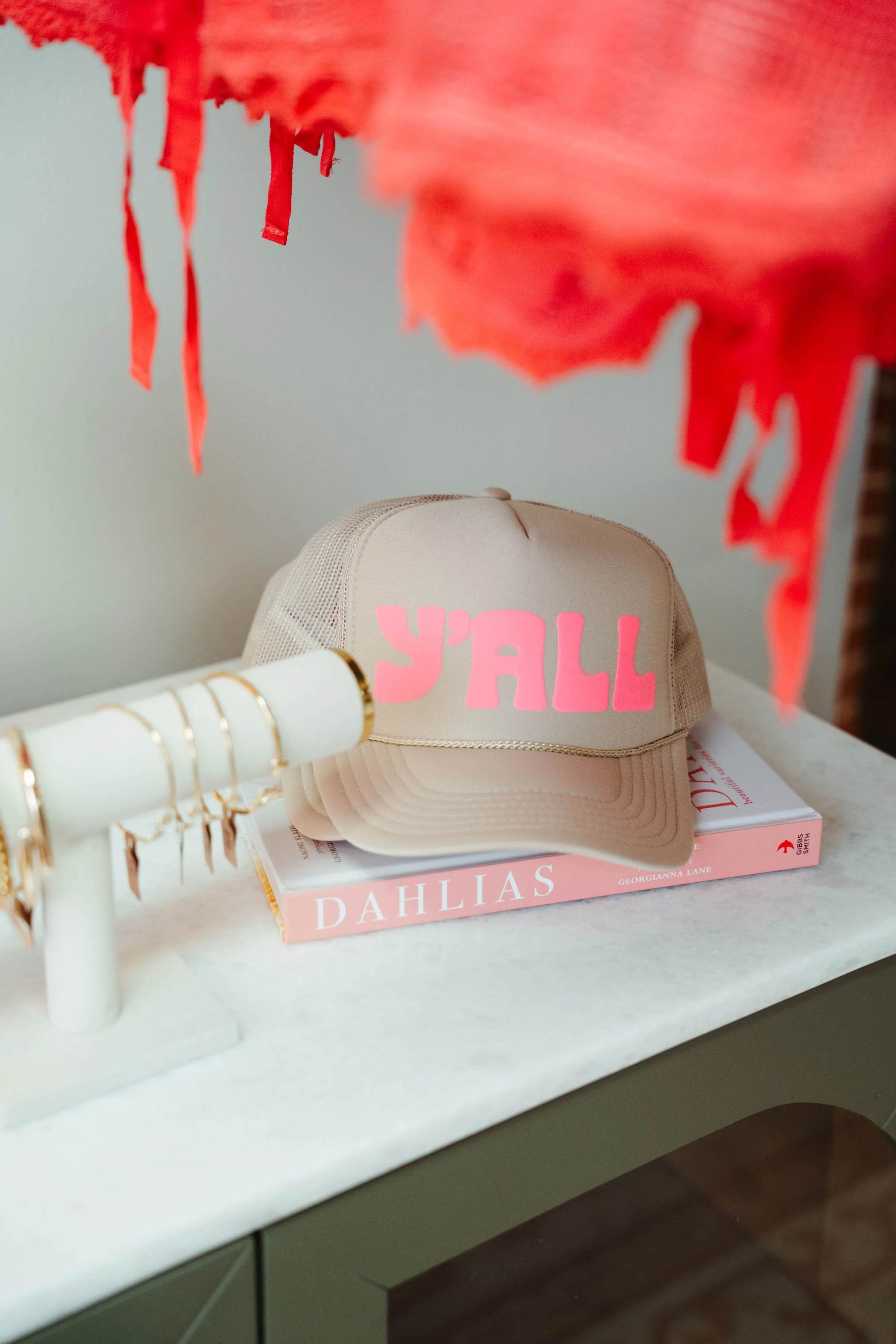 A beige baseball cap with pink letters that spell 'Y'ALL' on the front, placed on top of a pink book titled 'DAHLIAS' on a white surface. Part of a red decorative item with hanging strips is visible at the top of the image.
