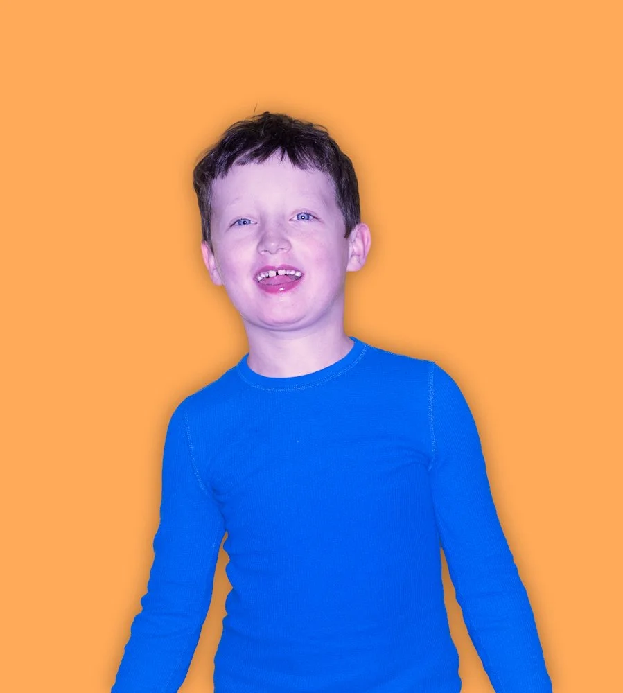 A young boy with short dark hair and blue eyes wearing a bright blue long-sleeve shirt, standing in front of an orange background, smiling with his mouth slightly open.