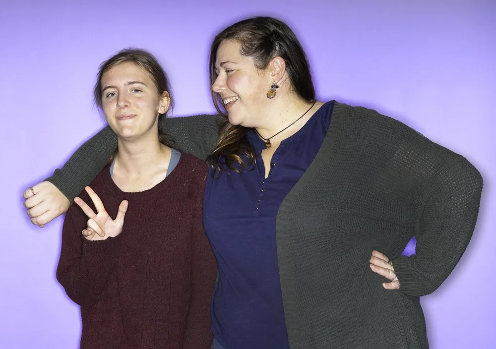 Young girl and woman smiling, with the girl's arm around the woman's shoulders, both displaying casual poses against a purple background.