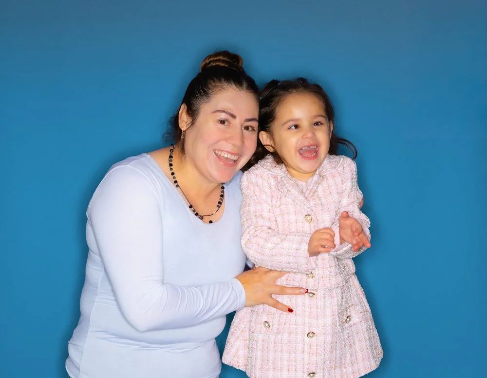 A woman and a young girl are smiling and laughing together against a blue background. The woman has dark hair in a bun, wears a white long-sleeve top, and a black necklace. The girl has dark hair, wears a pink and white textured coat, and is clapping her hands.