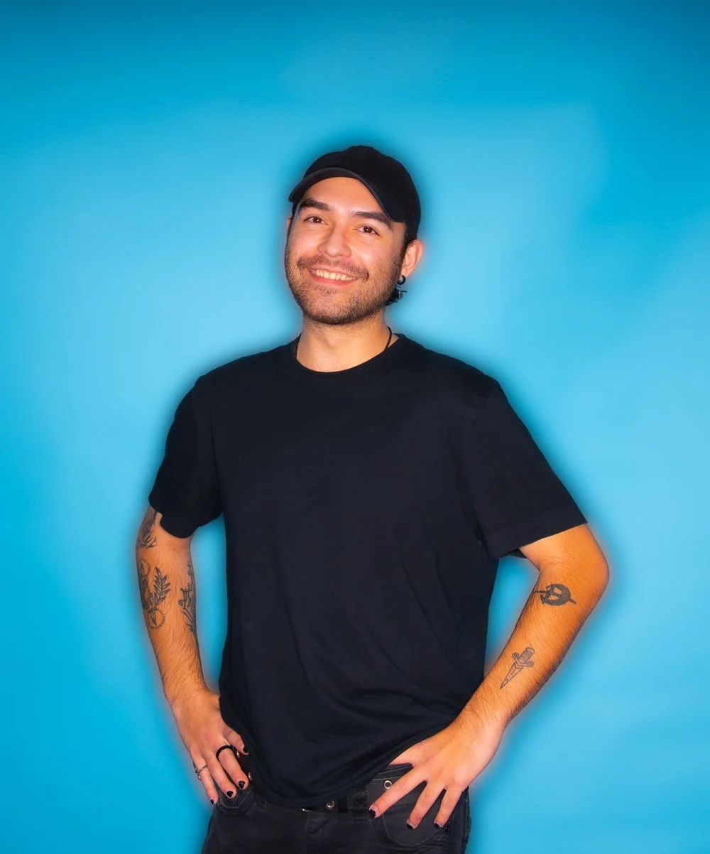 A young man with tattoos on his arms, wearing a black t-shirt and a black cap, smiling and posing with his hands on his hips against a blue background.