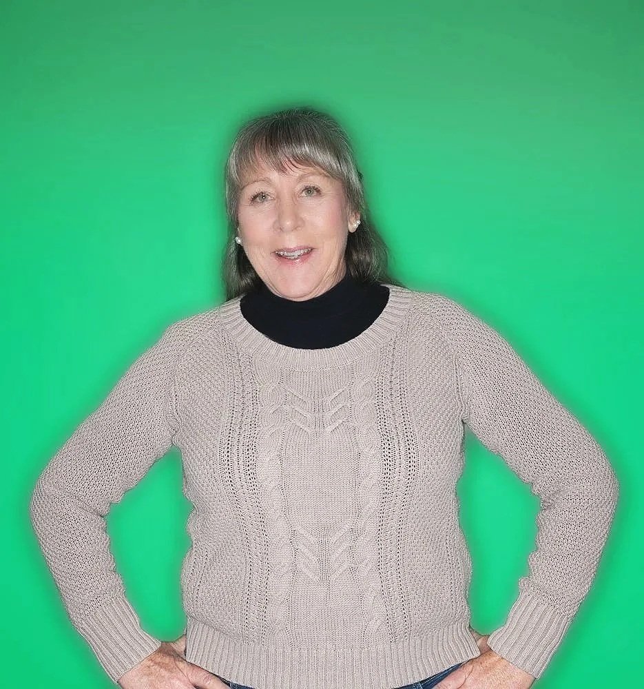 Woman with shoulder-length gray hair, wearing a light beige knitted sweater over a black turtleneck, standing with hands on hips against a green background.