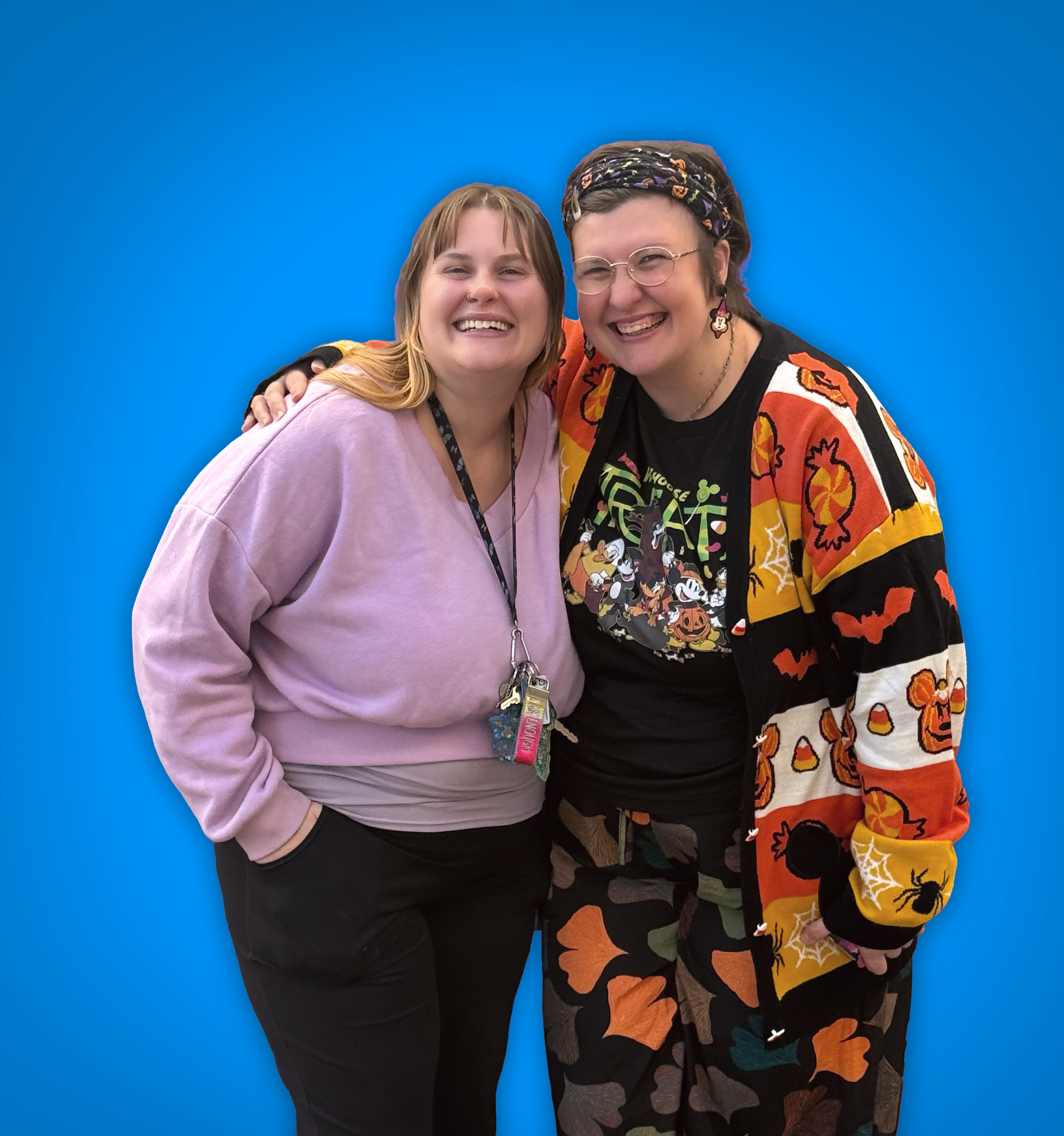 Two women smiling and standing close together against a blue background, one wearing a pink hoodie and the other wearing festive Halloween-themed clothing with a cardigan, t-shirt, and camouflage pants.