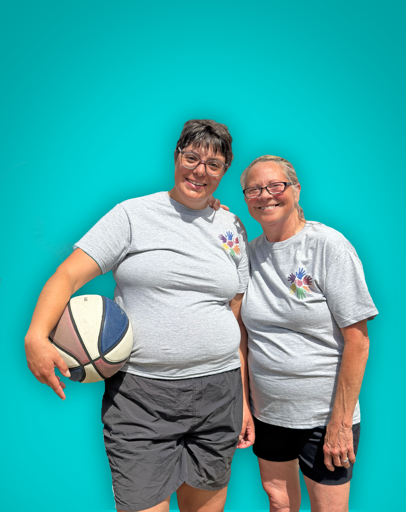 Two women smiling, wearing matching gray T-shirts with a colorful handprint logo, standing against a teal background. One woman is holding a basketball.