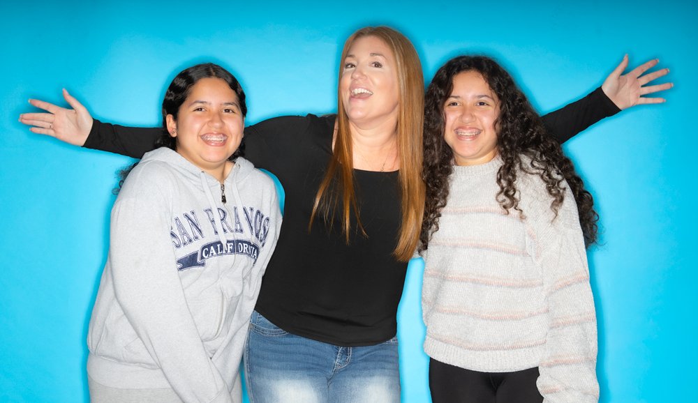 Three women smiling and posing together against a blue background, with the woman in the middle raising her arms.