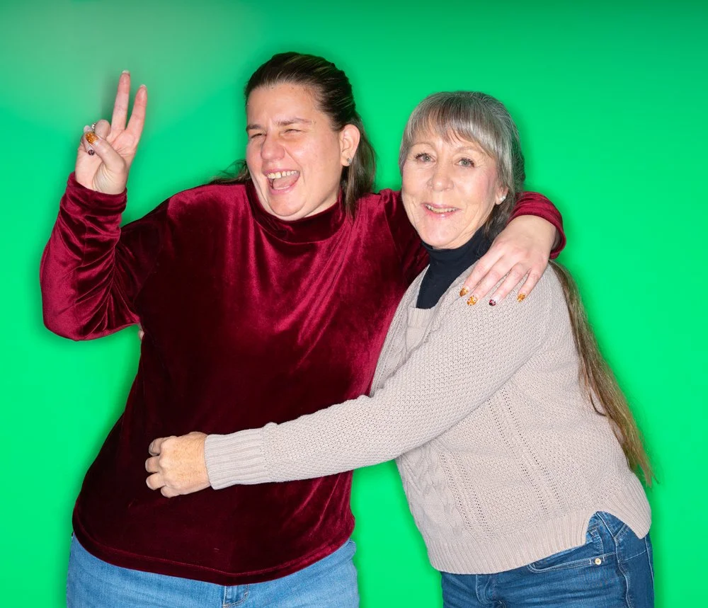 Two women hugging and smiling at the camera against a green background, one showing a peace sign with her left hand.