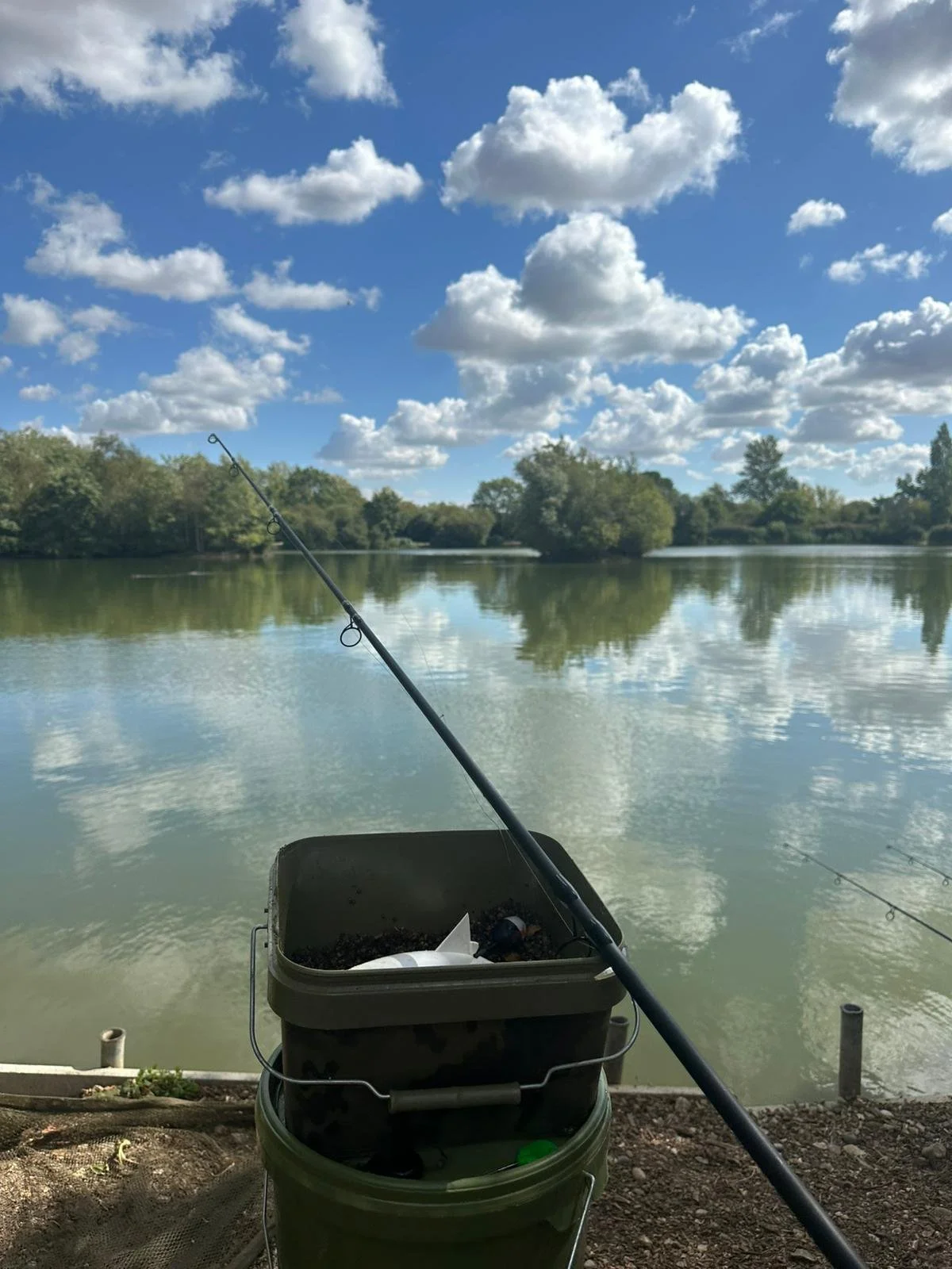 Fishing rod propped over a bucket on the shore of a lake with trees and a partly cloudy sky reflected in the water.
