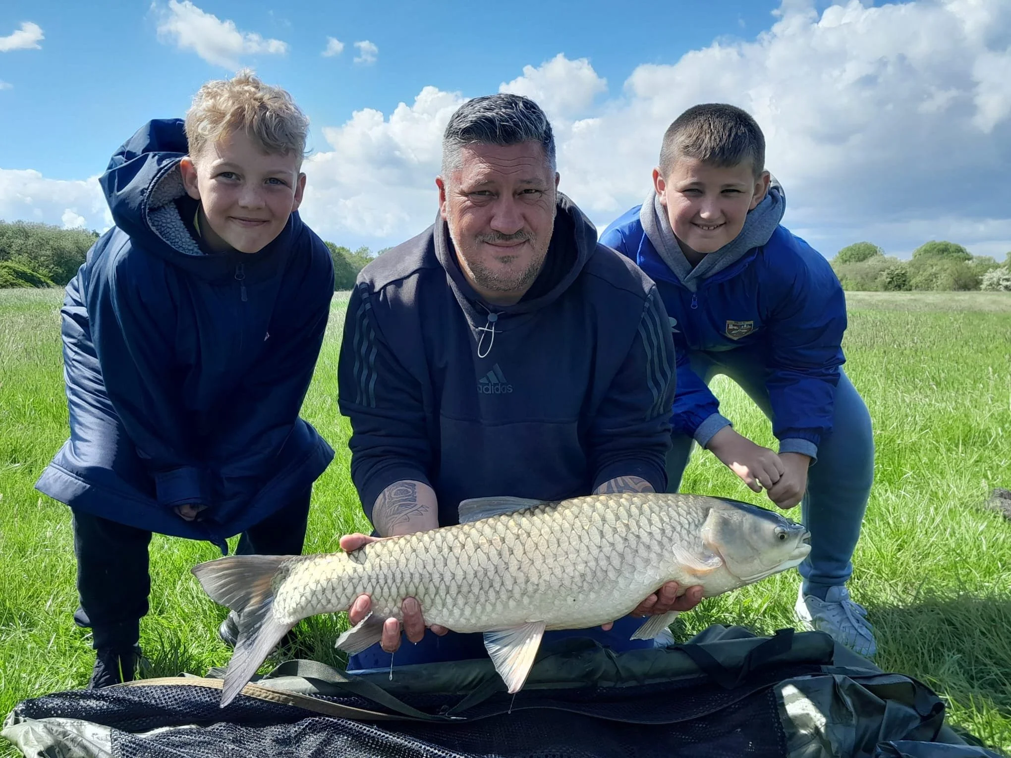 People at an outdoor camping site with a tent, trees, a person pushing a wagon, and another person walking away, under a partly cloudy sky.