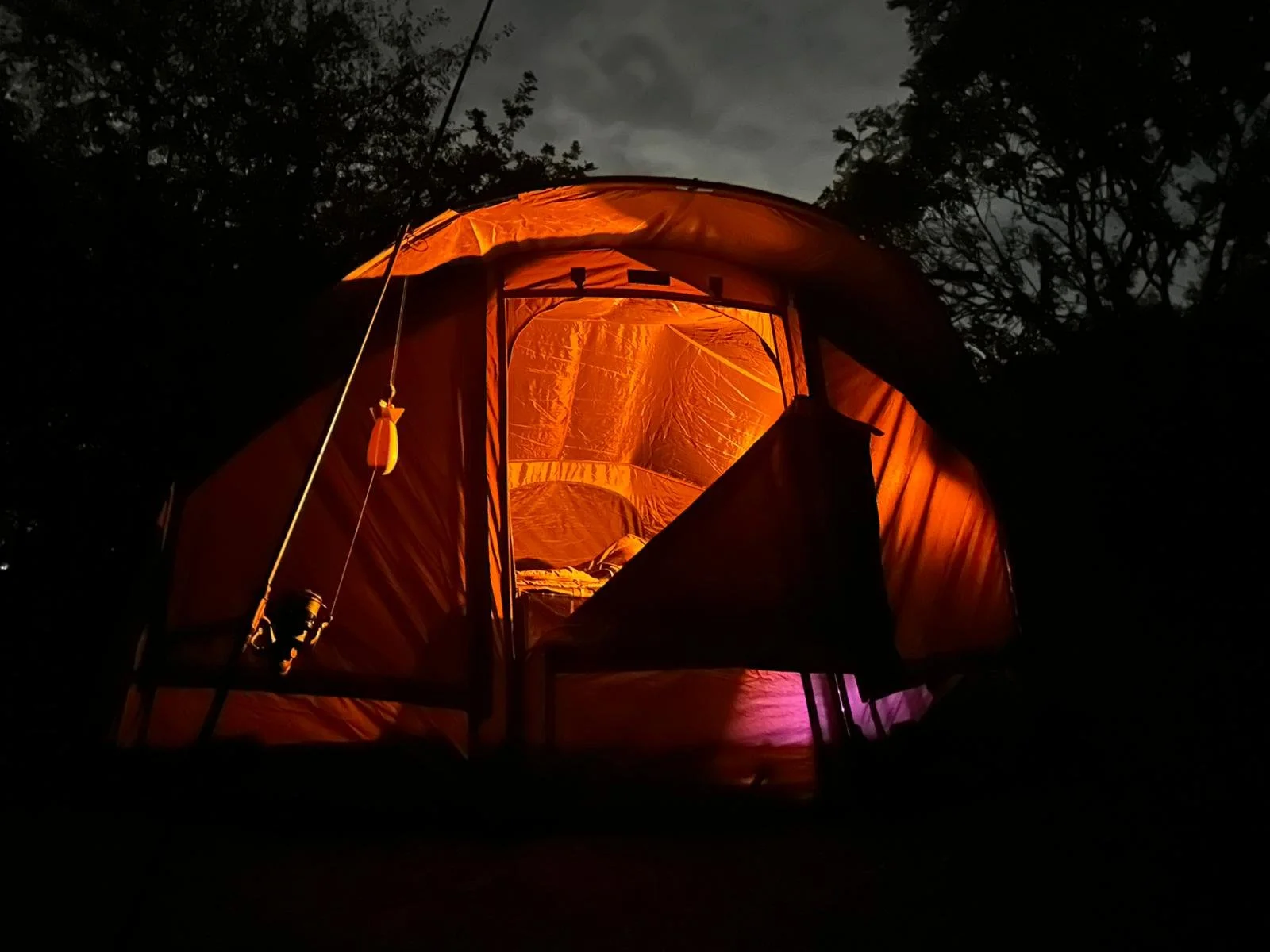 An illuminated orange tent set up outdoors at night, with trees and a cloudy sky in the background.