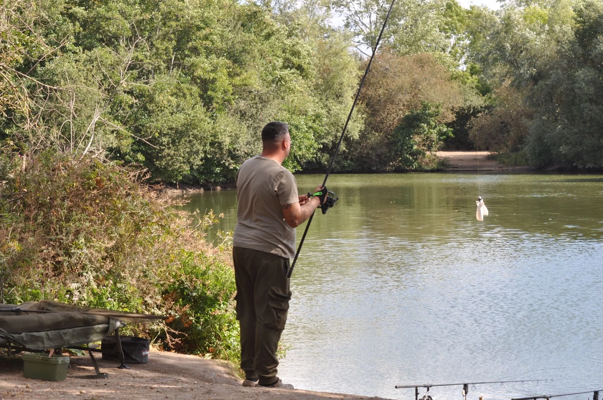 A man fishing by a river with trees in the background, holding a fishing rod, with a fish caught on the line.