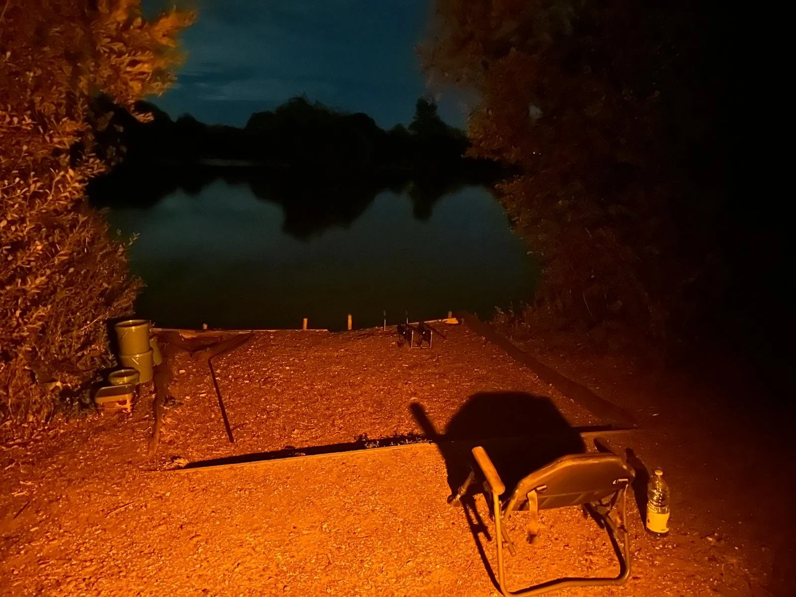 A nighttime scene at a lakeside with a camping chair, a bottle of water, and two trash cans on a gravel path illuminated by an orange light, with trees framing the water and a dark sky overhead.
