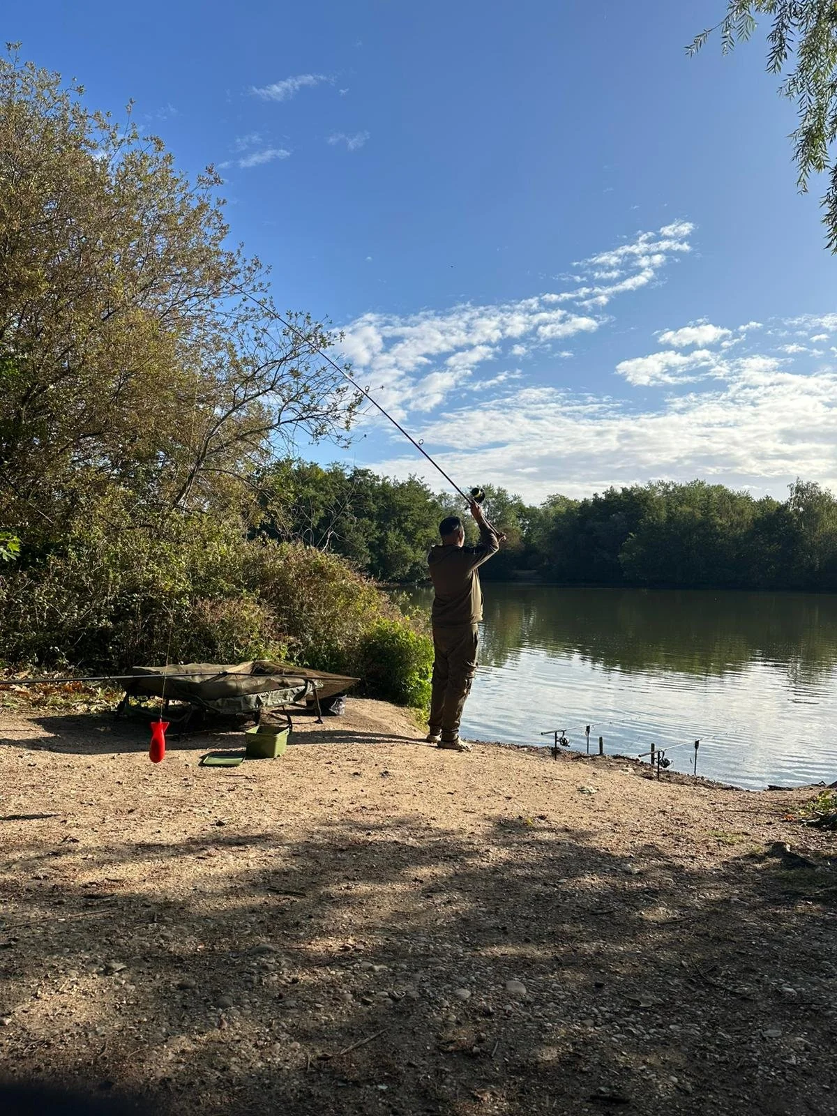 Person fishing by a river on a sunny day with a blue sky and scattered clouds, surrounded by trees.