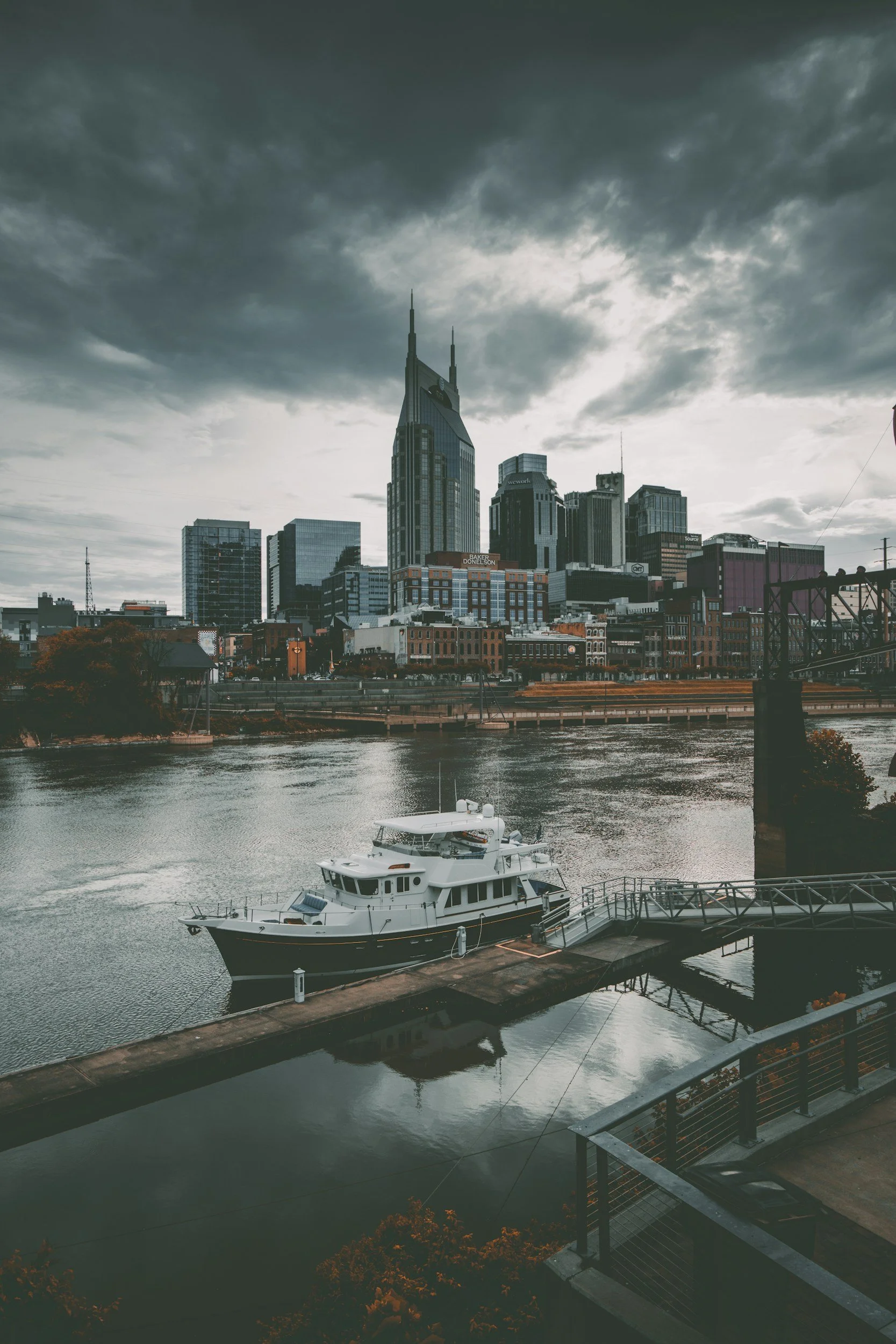 A city skyline with tall buildings under a dark, cloudy sky, a river with a white yacht docked at a pier in the foreground.