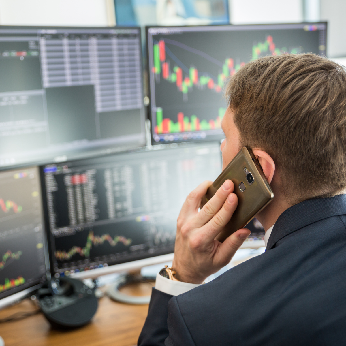 Man in business suit with short brown hair using a cellphone while monitoring multiple computer screens displaying financial trading charts and data.