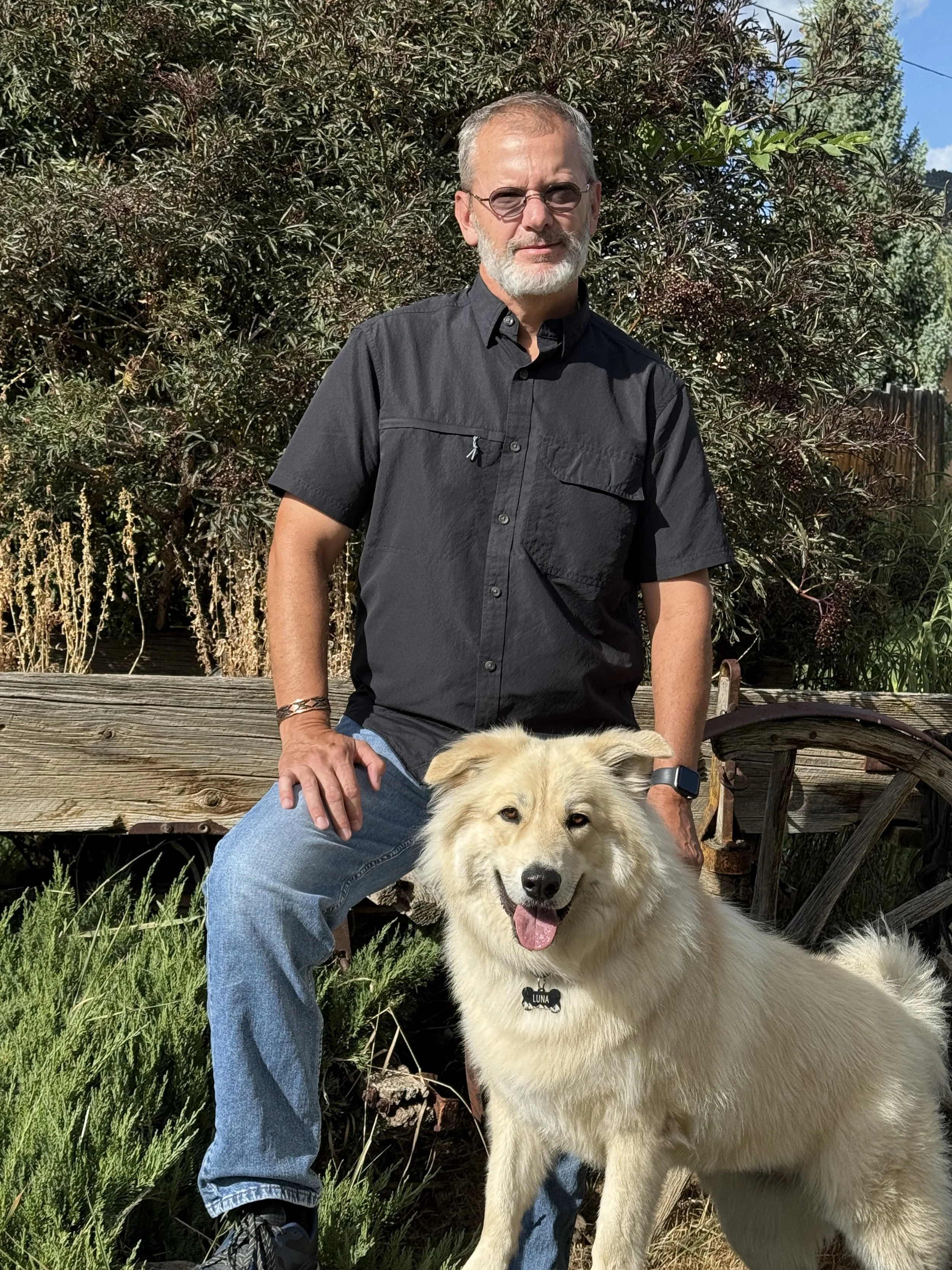 A man with gray hair, glasses, and a beard, wearing a black short-sleeve shirt and jeans, is sitting outdoors on a wooden bench with a happy, light-colored dog wearing a collar with a name tag that reads Luna. They are in a garden with green plants and a wooden fence.