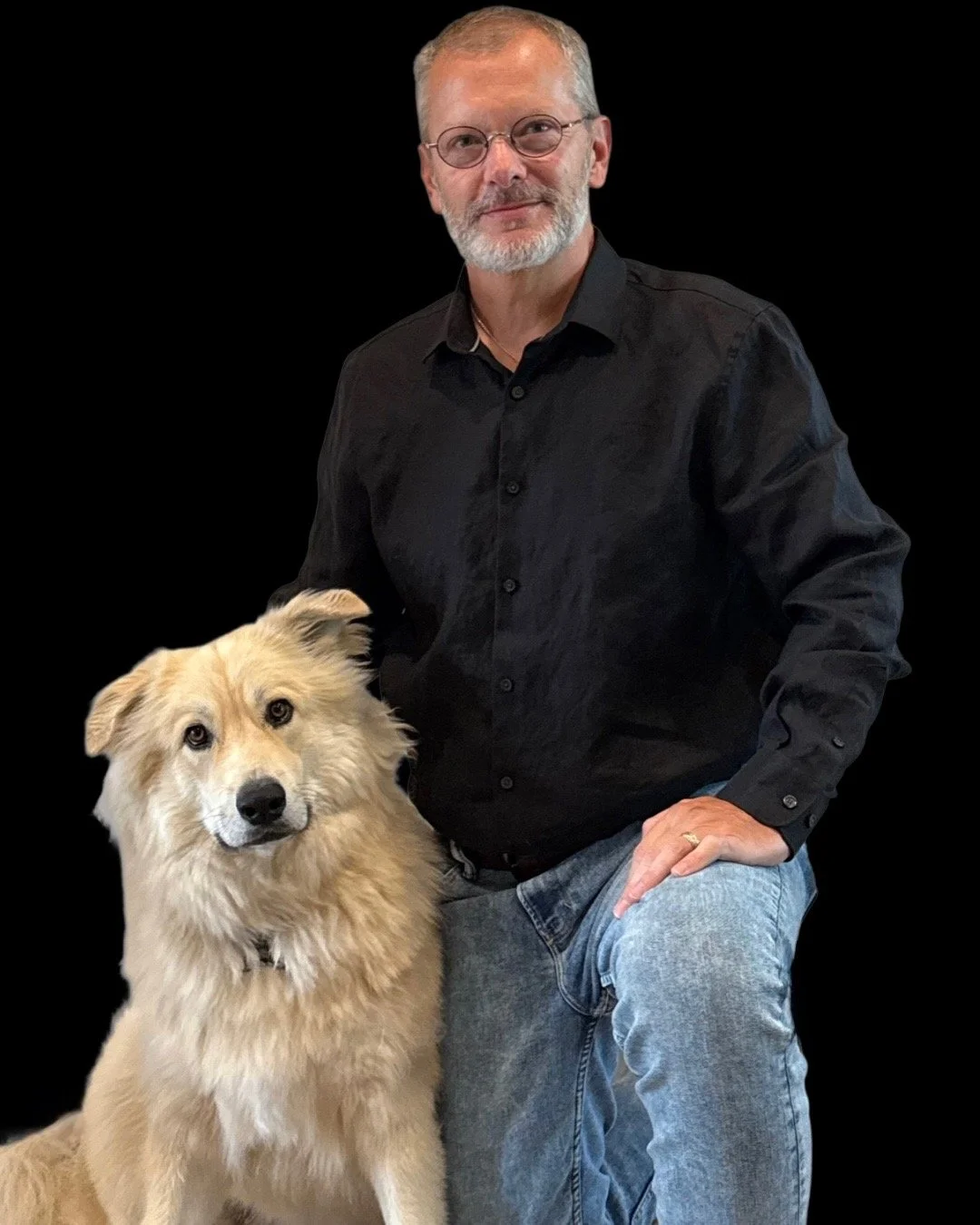 A man with gray hair, glasses, and a beard wearing a black shirt, kneeling next to a golden retriever with a black background.