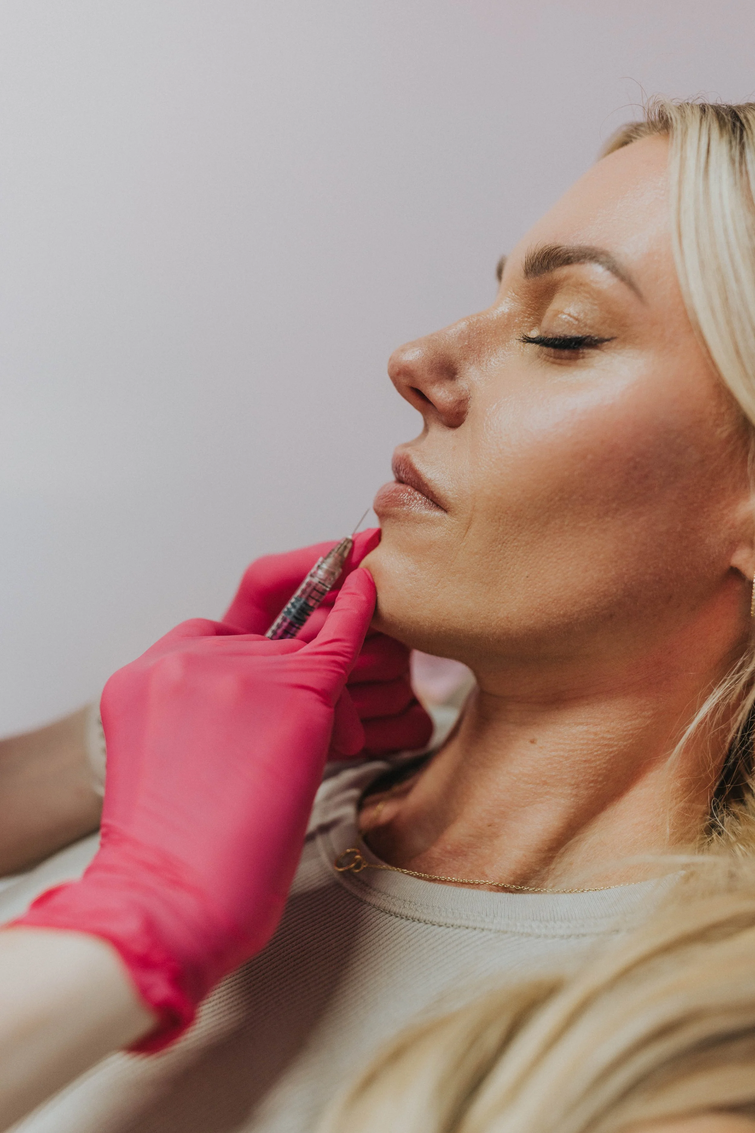 A woman with blonde hair receives a cosmetic injection in her chin area from a medical professional wearing pink gloves.