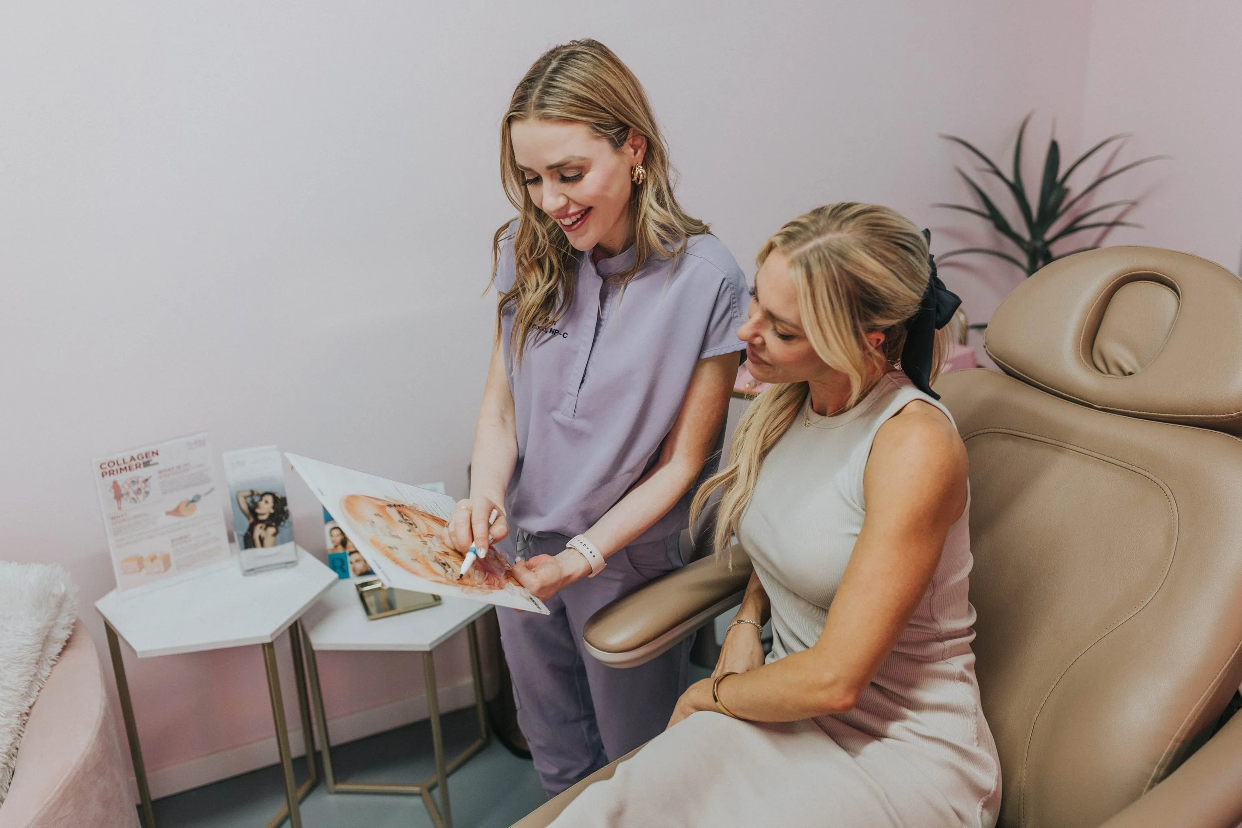 A woman in medical scrubs showing a brochure to another woman in a beige dress in a spa or cosmetic clinic.