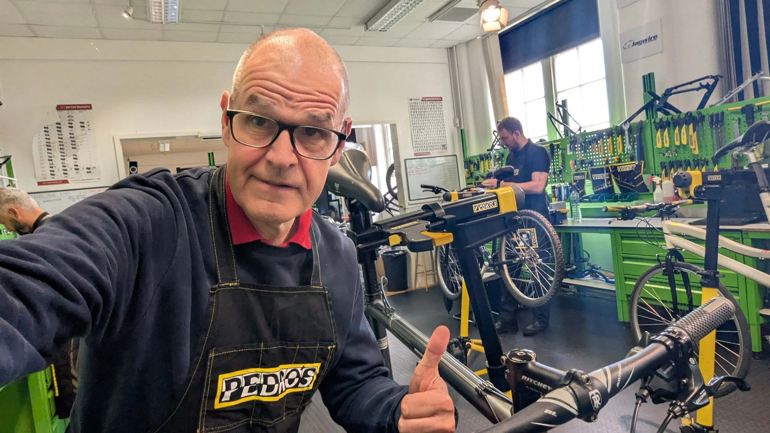 A man wearing glasses, a black Pedros apron, and a red shirt taking a selfie inside a bike shop filled with bicycle tools and bikes.