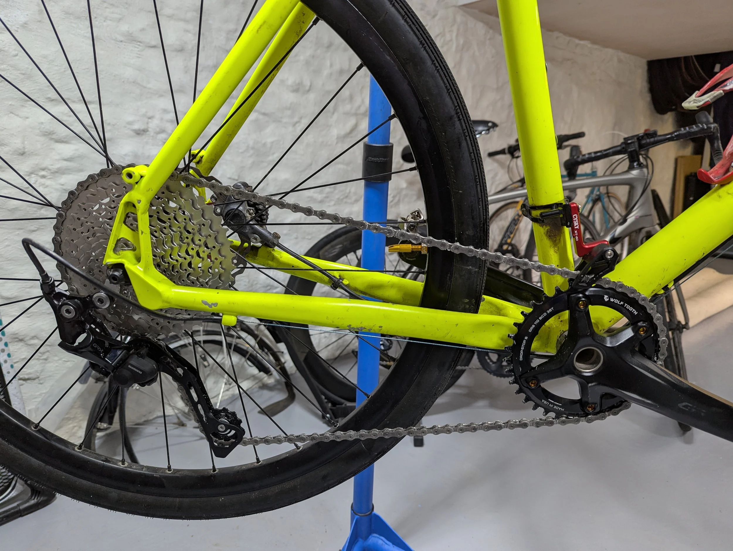 Close-up of a yellow mountain bike with a black crankset and derailleur, mounted on a blue repair stand, with other bikes in the background.