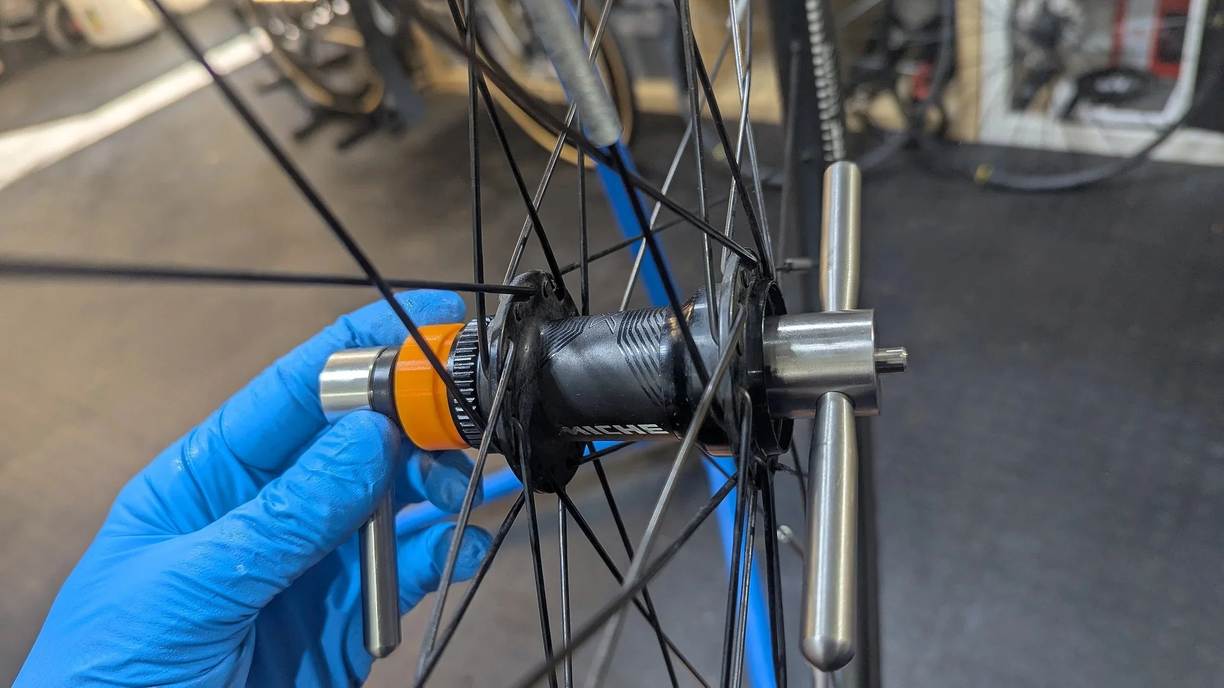 A person's gloved hand holding a bicycle hub with orange and silver parts, close to a wheel with multiple spokes.