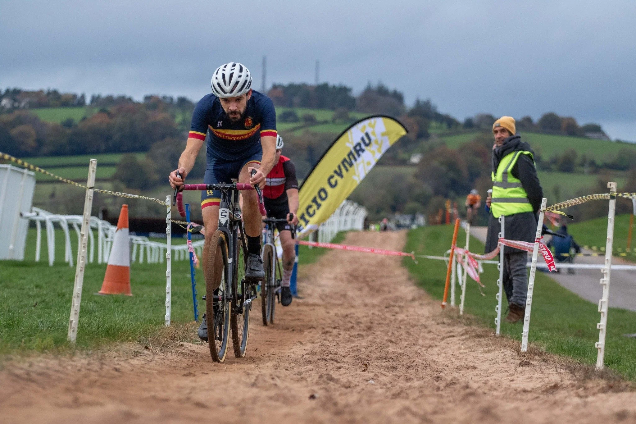Cyclists participating in a mountain biking race on a dirt trail, with a race official standing on the side, greenery in the background, and cloudy skies overhead.