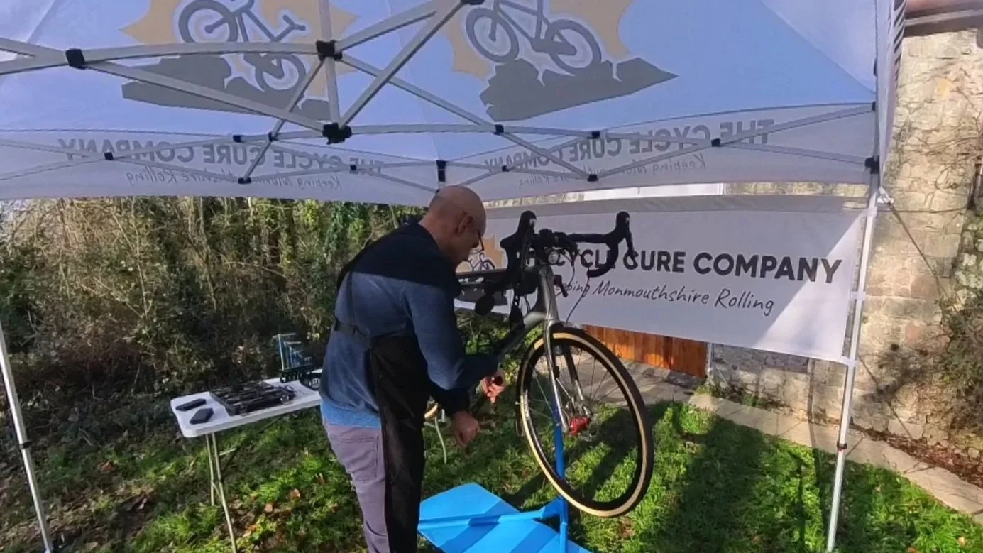 A mechanic working on a bicycle under a canopy with the logo and name of a bicycle repair company, outside near a stone wall and bushes.