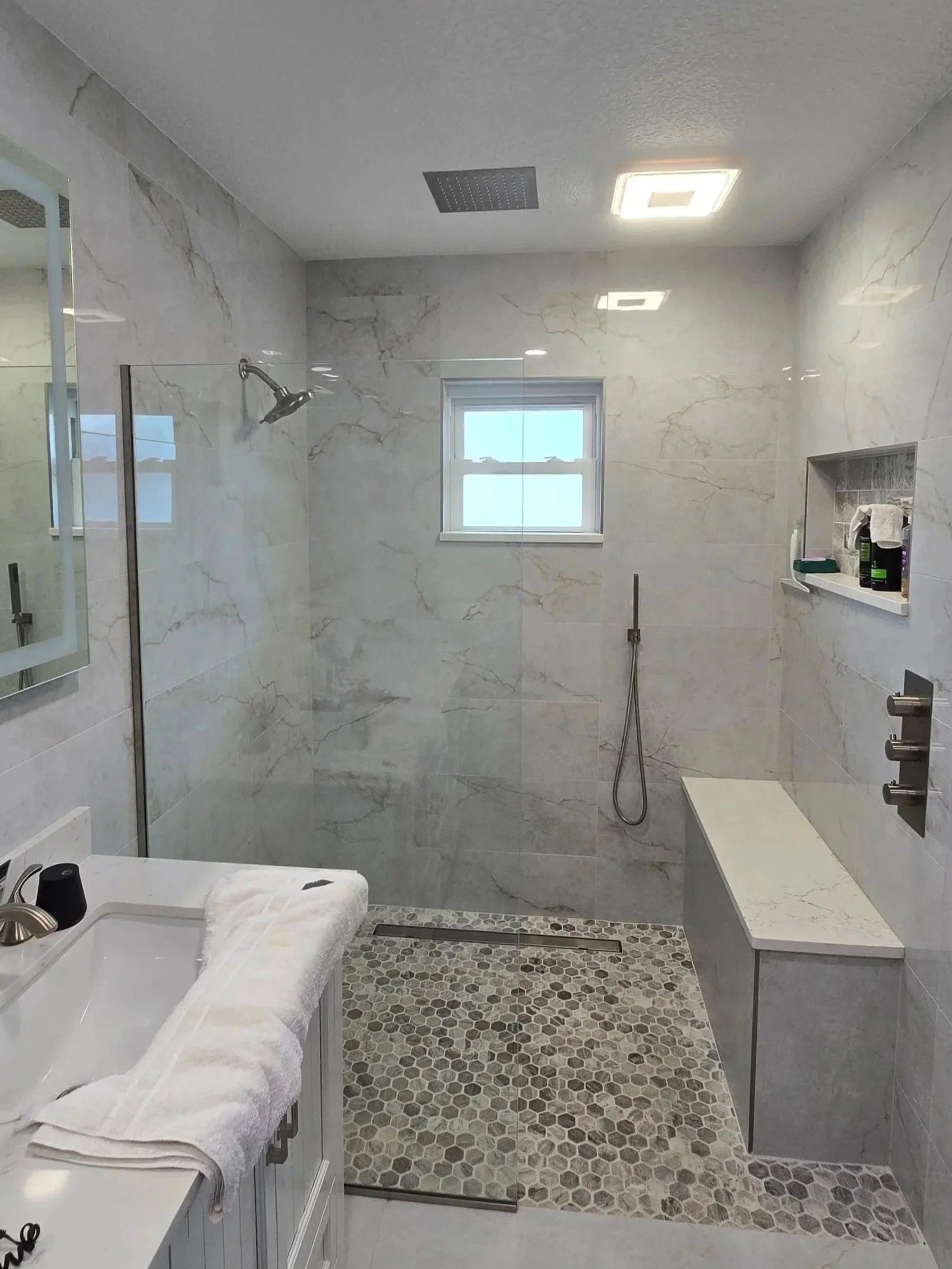 Modern bathroom with marble walls, walk-in shower with glass door, small window, pebble tile floor in shower, built-in wall niche with toiletries, white vanity with towels, mirror, and hanging towels