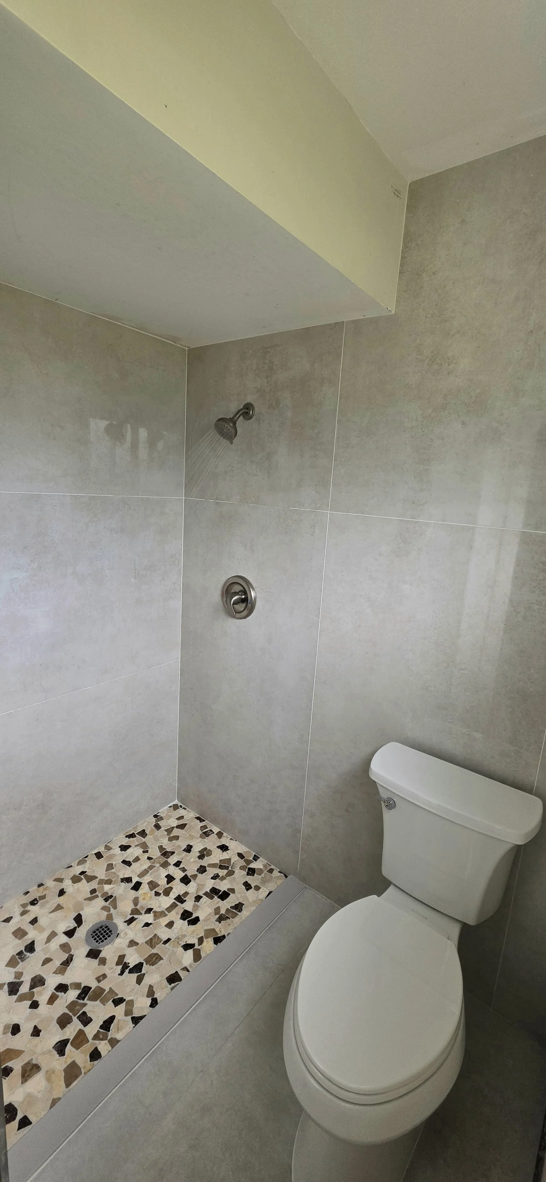 Bathroom with a shower area that has a wall-mounted showerhead, a floor drain with a pebble mosaic pattern, and a toilet next to the shower, all with gray tiled walls.