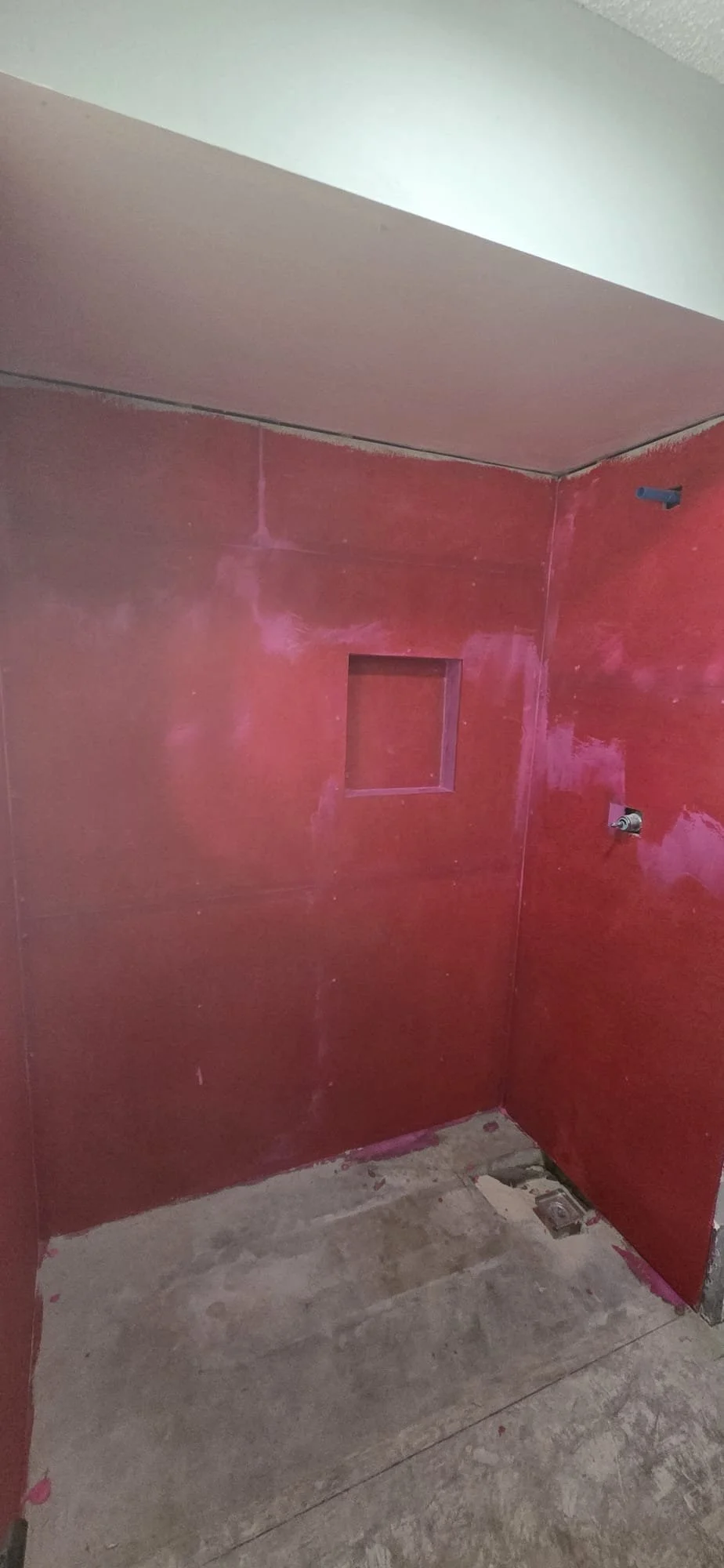 A bathroom under renovation with red painted walls, a small square cutout in the wall, a toilet flush connector, and a floor drain.