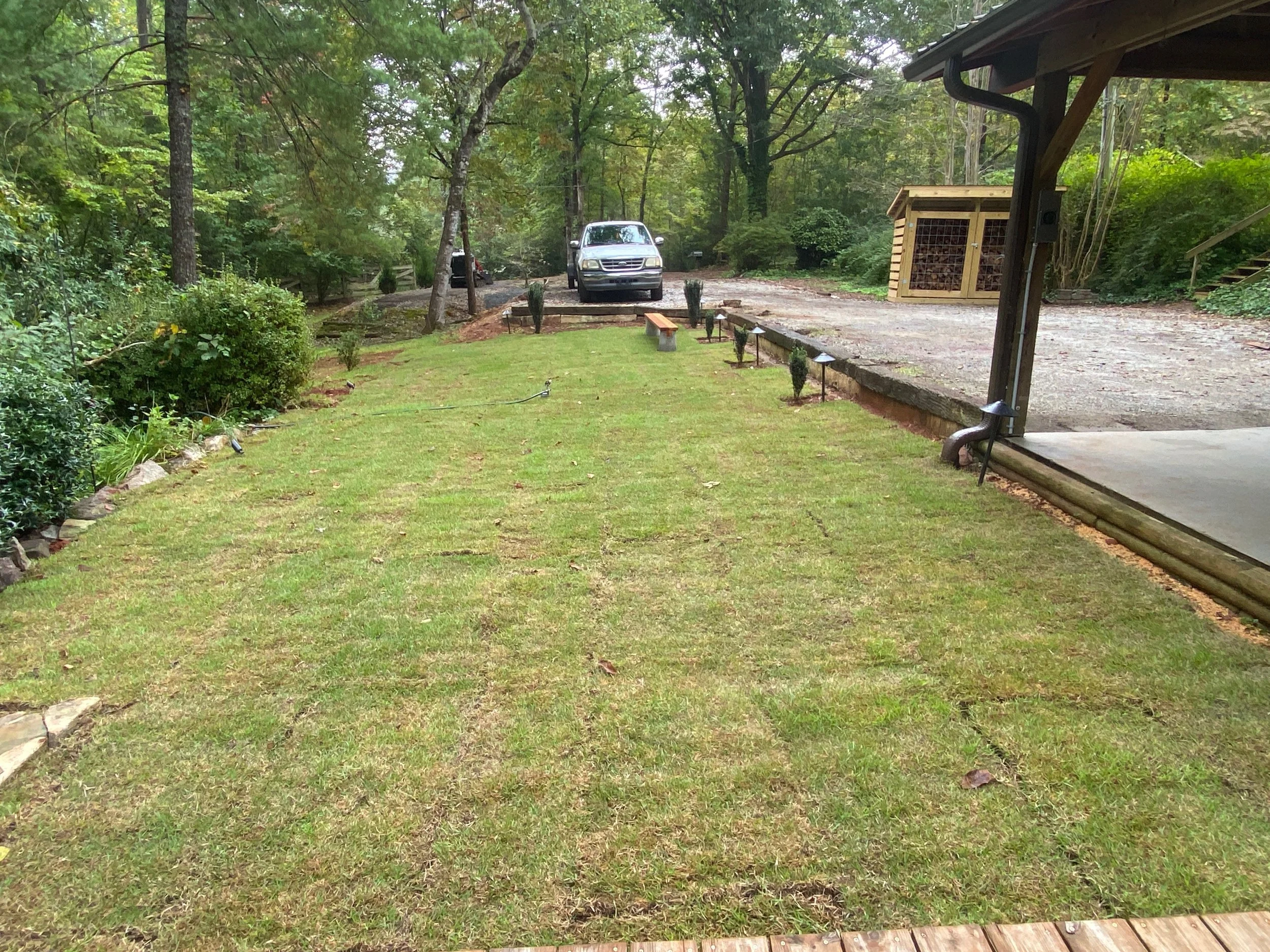 A backyard lawn area with grass, shrubs, trees, and a gravel driveway at the back with a parked car and a wooden shed. There are small garden lights along the lawn edge and a bench.