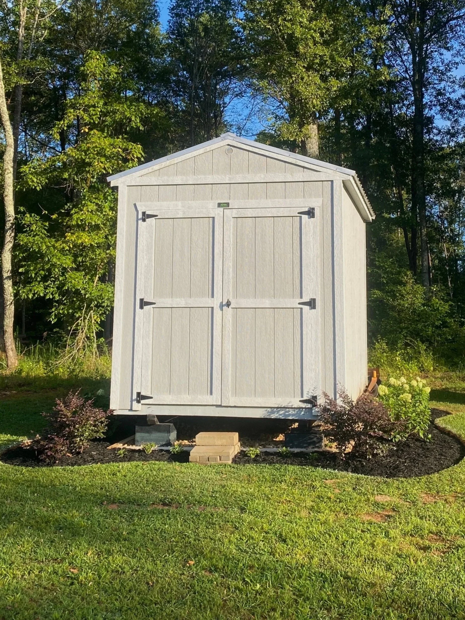 A small gray wooden shed with double doors, situated on a grassy area with small plants and bushes in front, surrounded by trees under a clear blue sky.