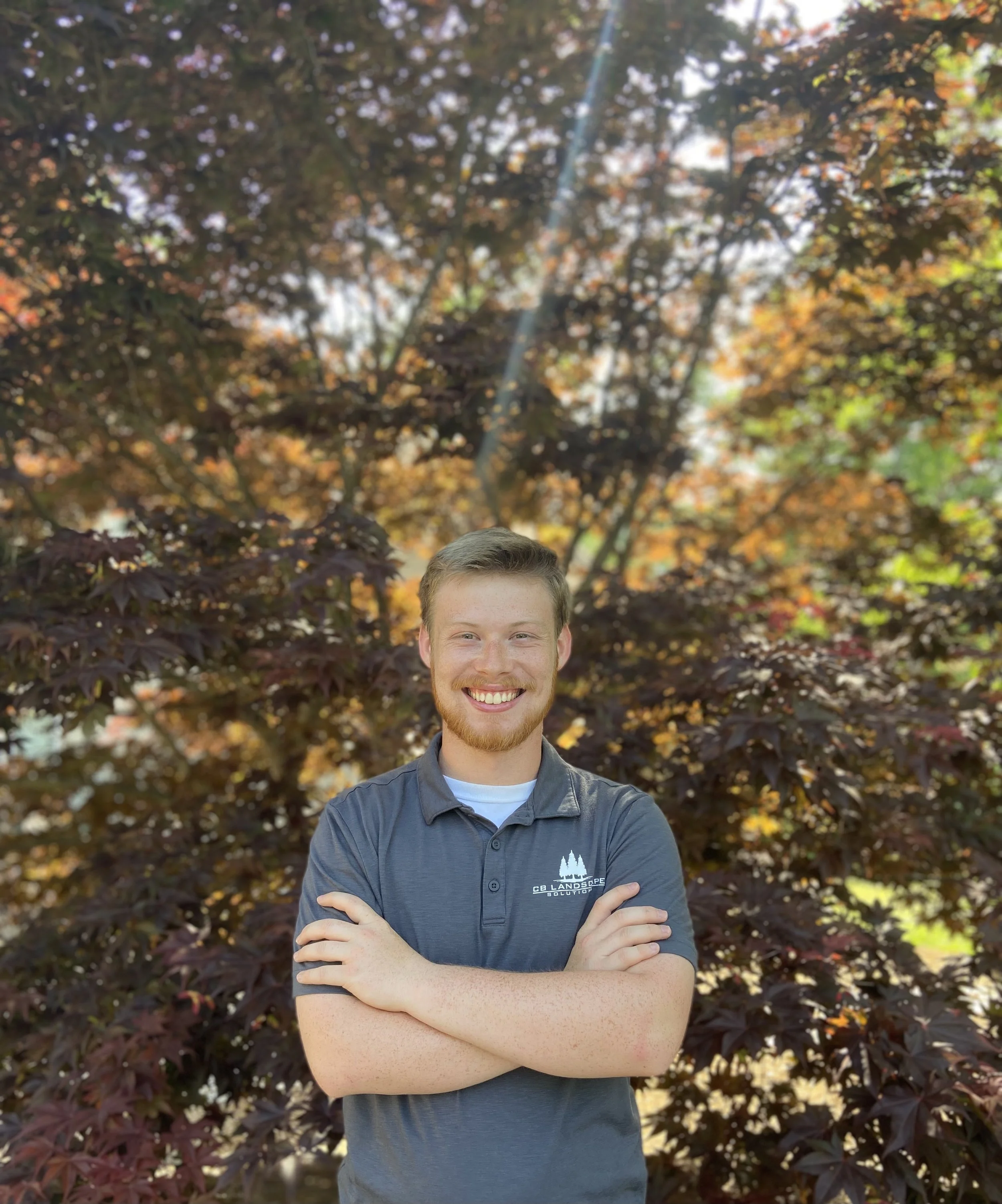 A man with light brown hair and a beard smiling, standing outdoors in front of autumnal trees with red and orange leaves, with his arms crossed.