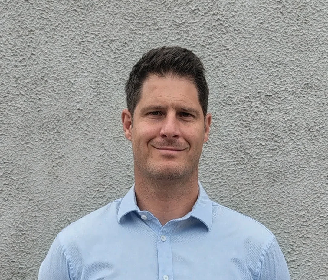 A man with dark hair and a light blue collared shirt standing against a textured gray wall.