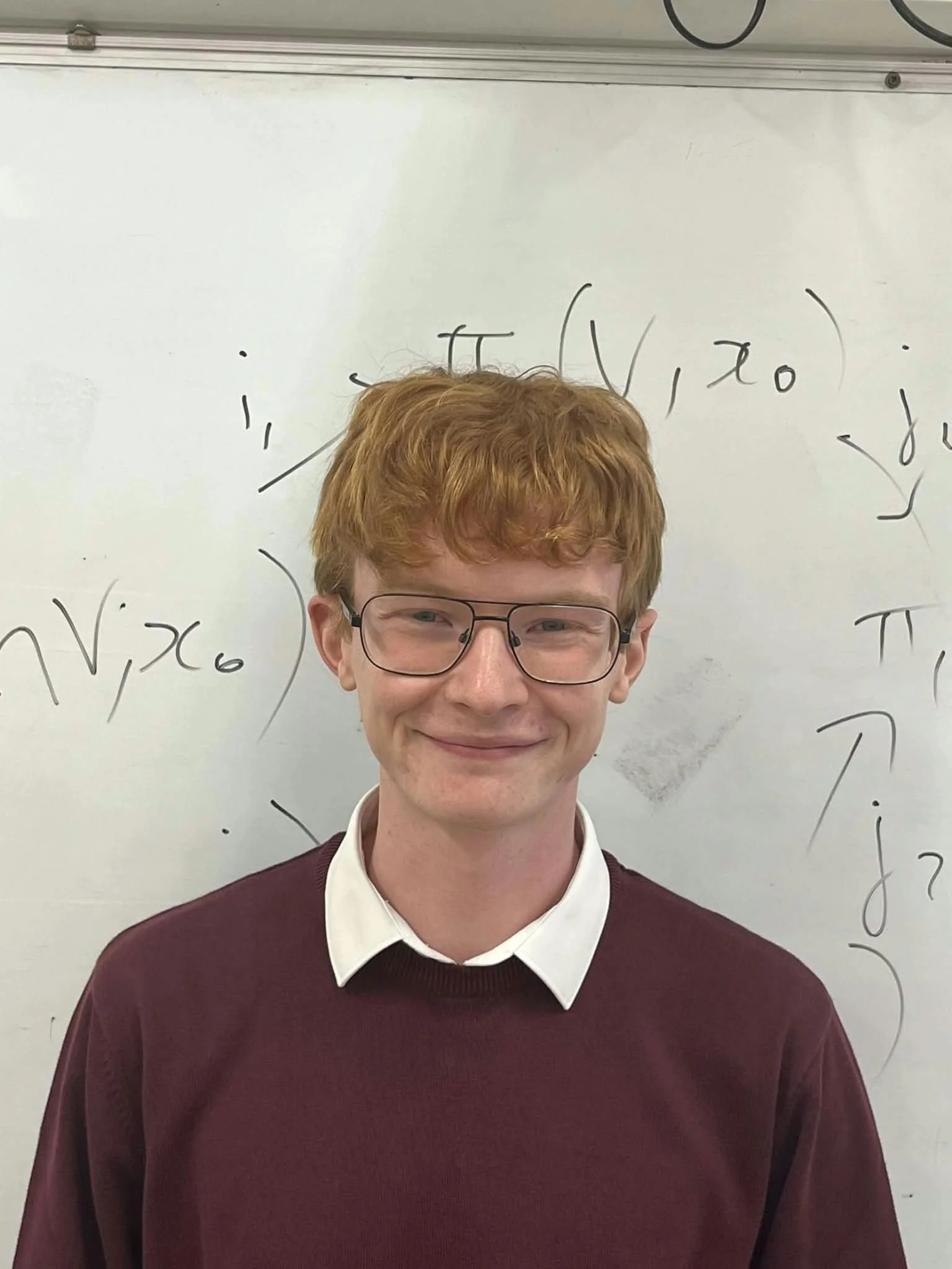 A young person with red hair, glasses, and fair skin, wearing a white collared shirt under a maroon sweater, smiling in front of a whiteboard with mathematical equations written on it.