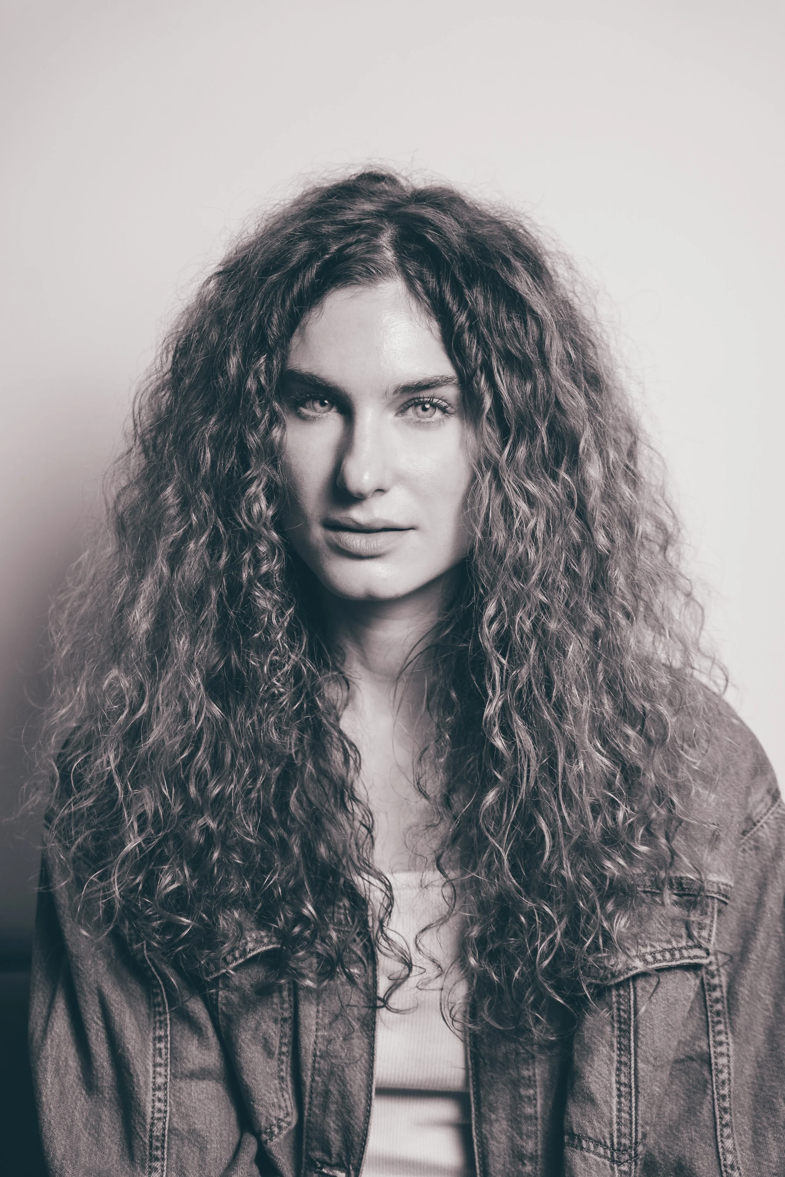 Black and white portrait of a woman with long, curly hair, wearing a denim jacket and a white top, looking directly at the camera.