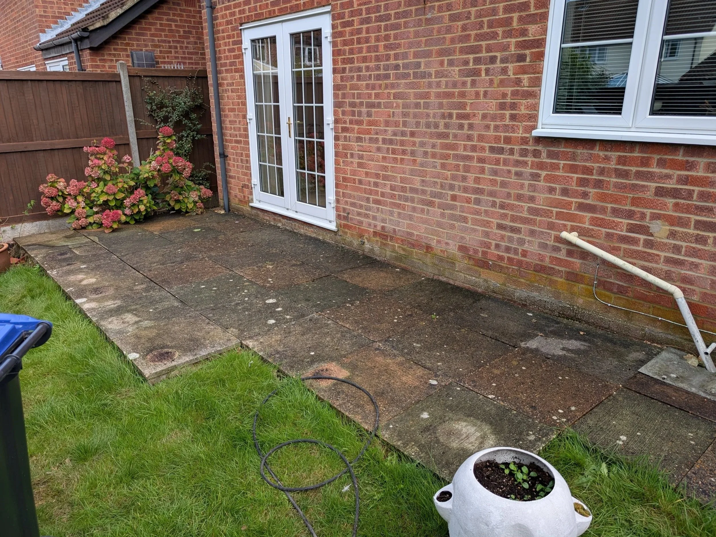 Backyard patio with stone slabs next to a brick house, gardening tools, and a flowering bush in the corner.