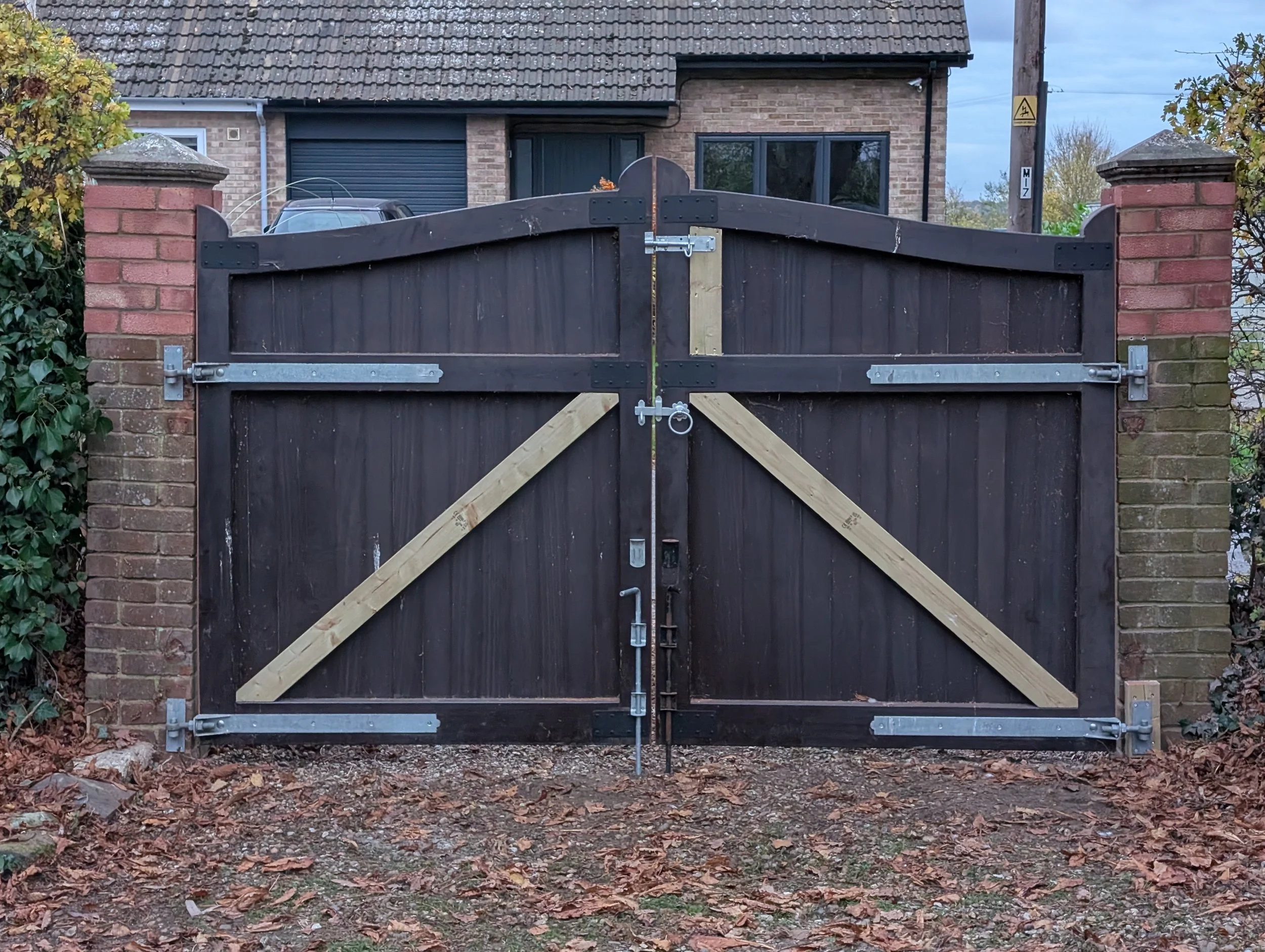 A pair of black wooden gates with metal hinges and a latch, framed by brick pillars and a brick house in the background.