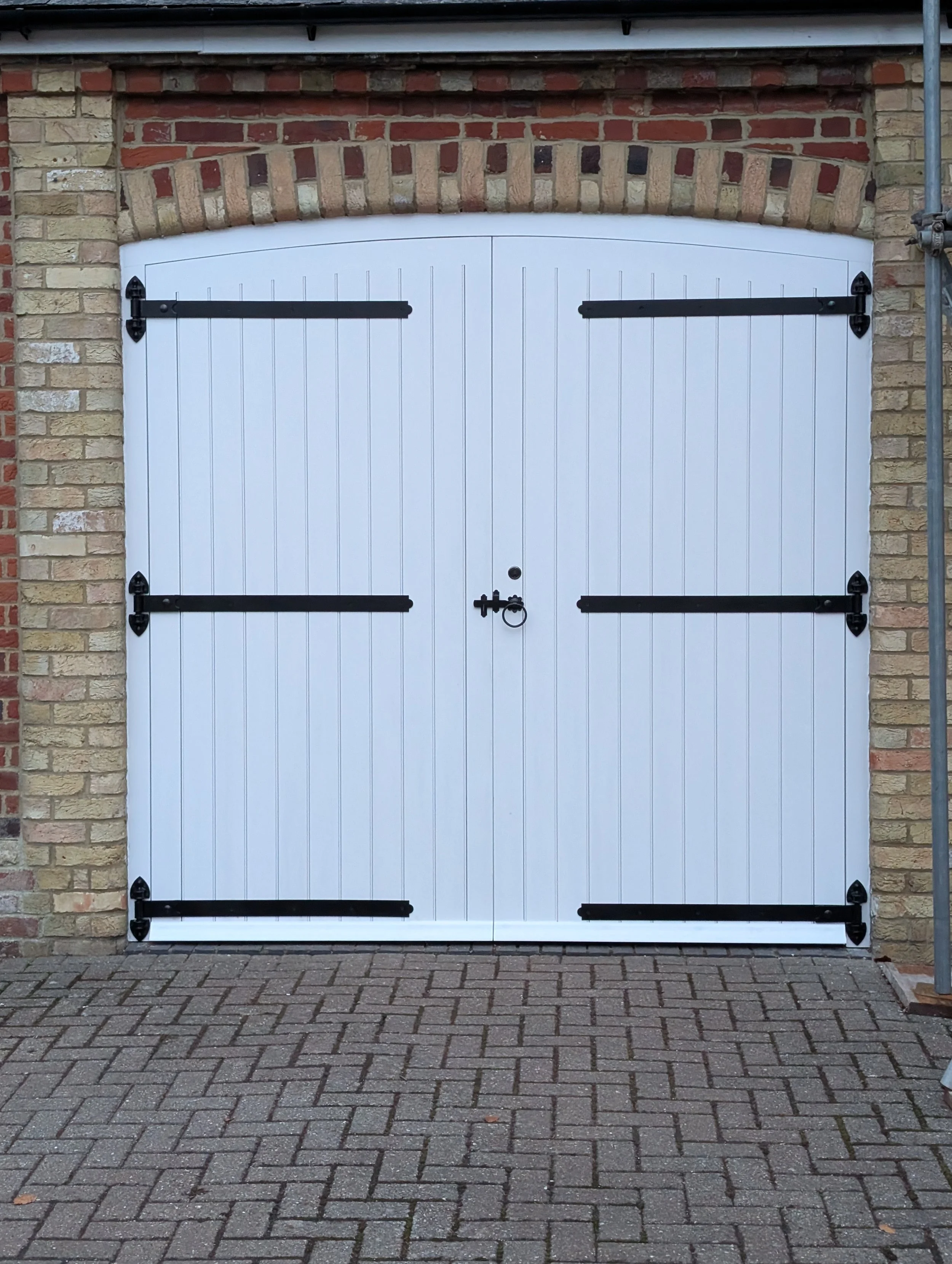 White garage door with black hinges and a black handle, set in a brick wall with a brick arch above, on a driveway with interlocking pavers.