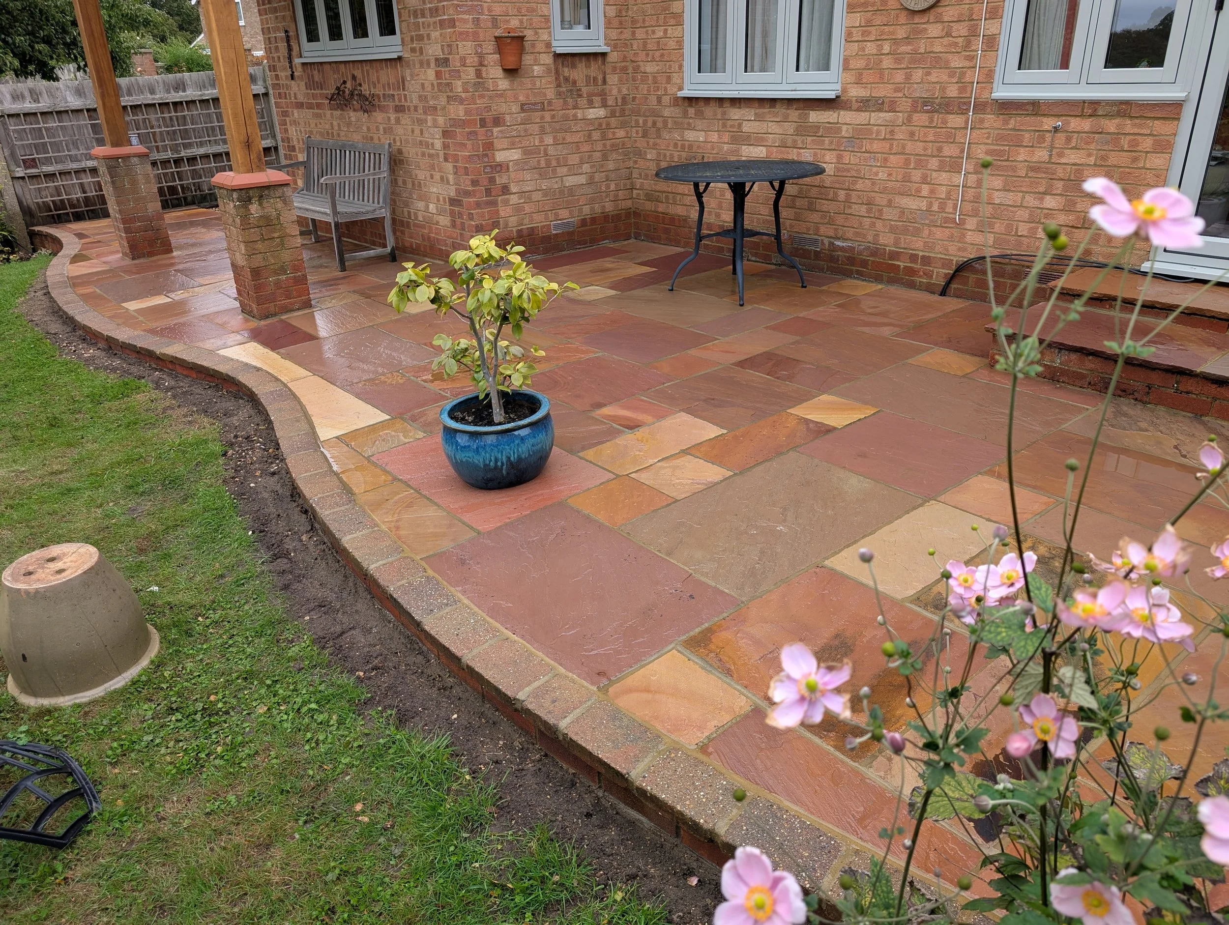 Newly paved patio with red and yellow sandstone tiles, decorated with potted plants, a bench, and a small round table, attached to a brick house with large windows.