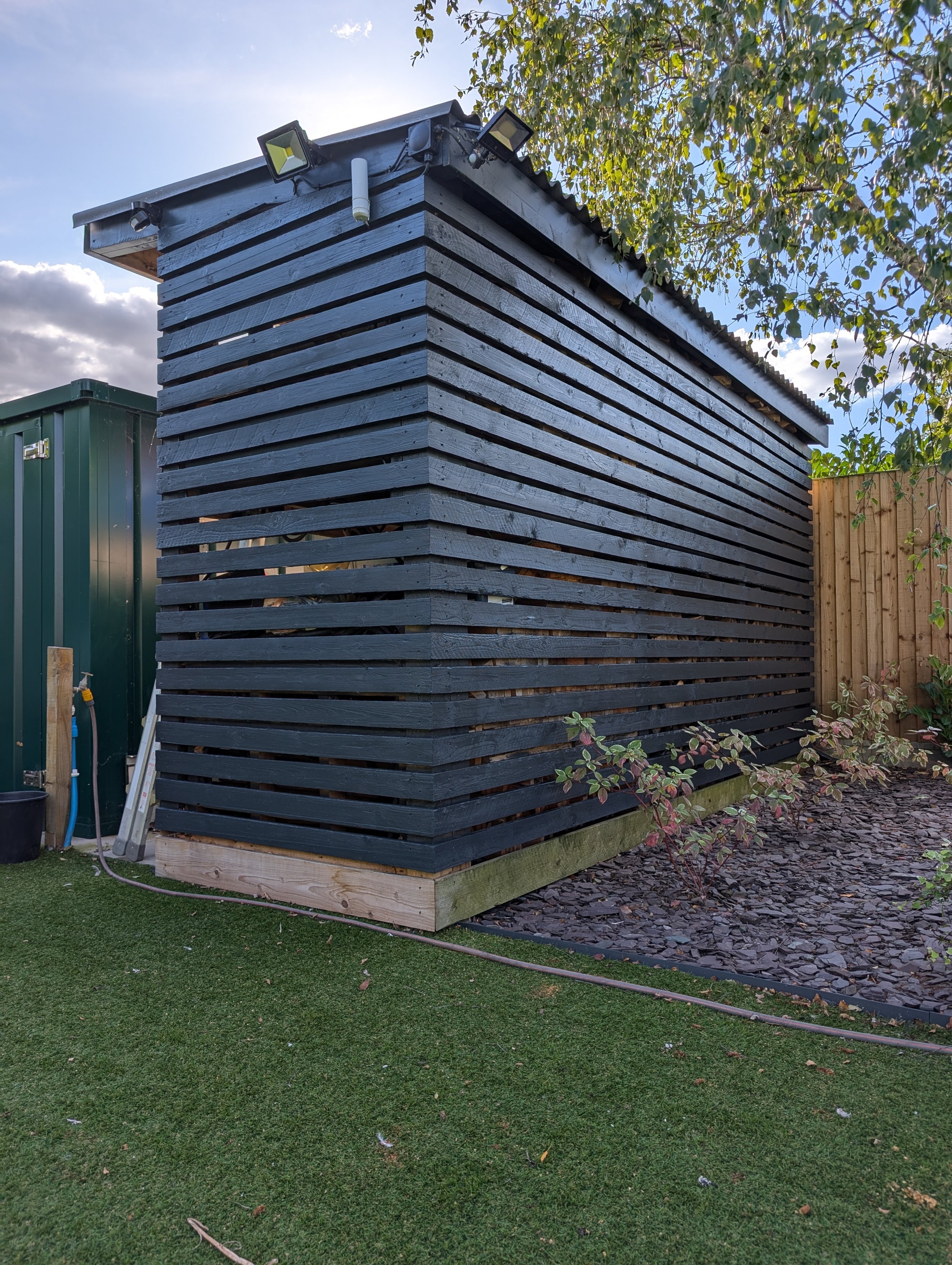 A small, rectangular wooden shed painted black with horizontal slats, situated on a grassy lawn with a wood-chip garden bed and a wood fence on the right, under a partly cloudy sky with trees nearby.