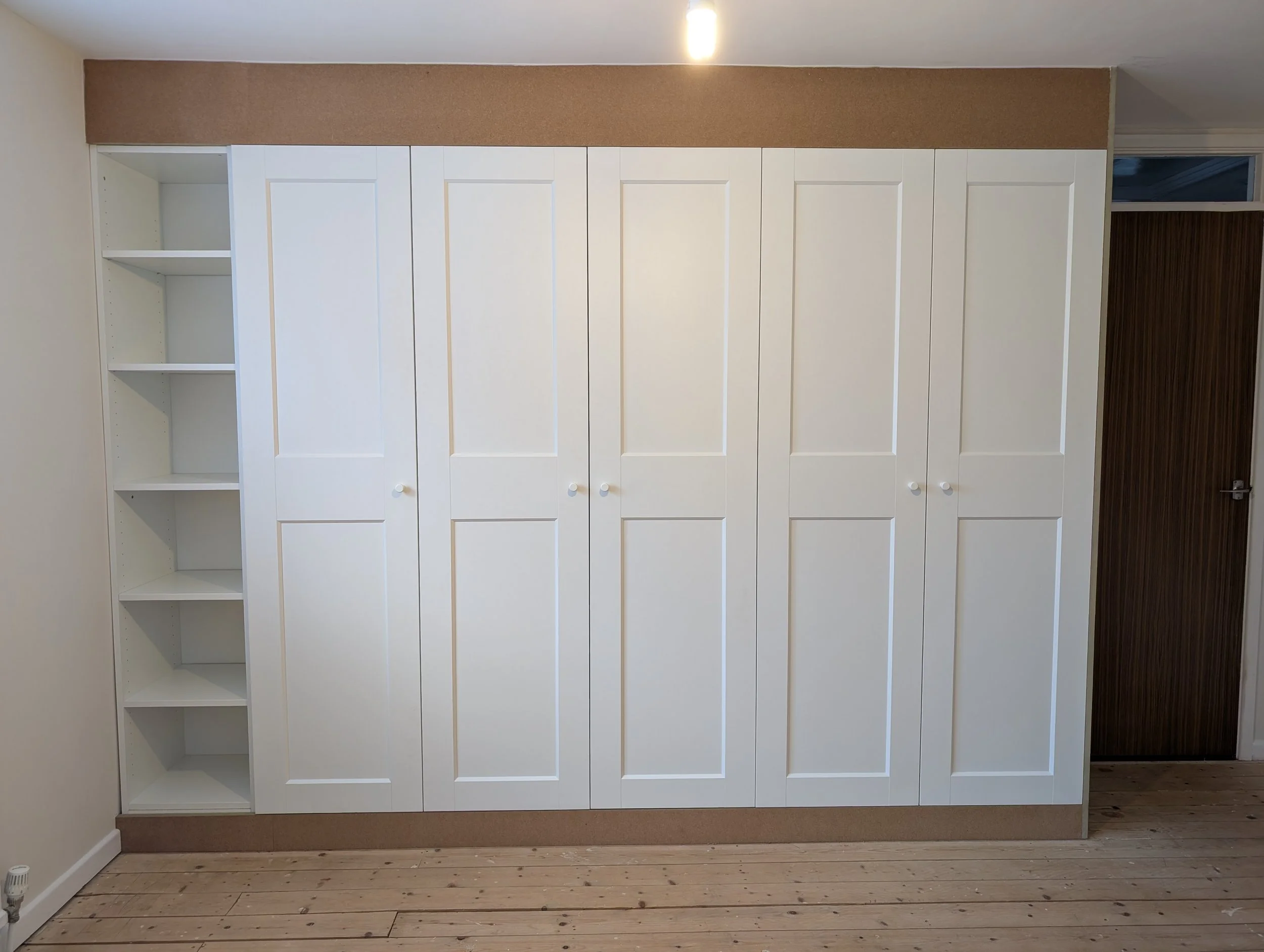 White built-in wardrobe with five doors next to an open shelving unit with five shelves. Brown trim at the top and bottom, on a wooden floor with a vent in the bottom left corner.