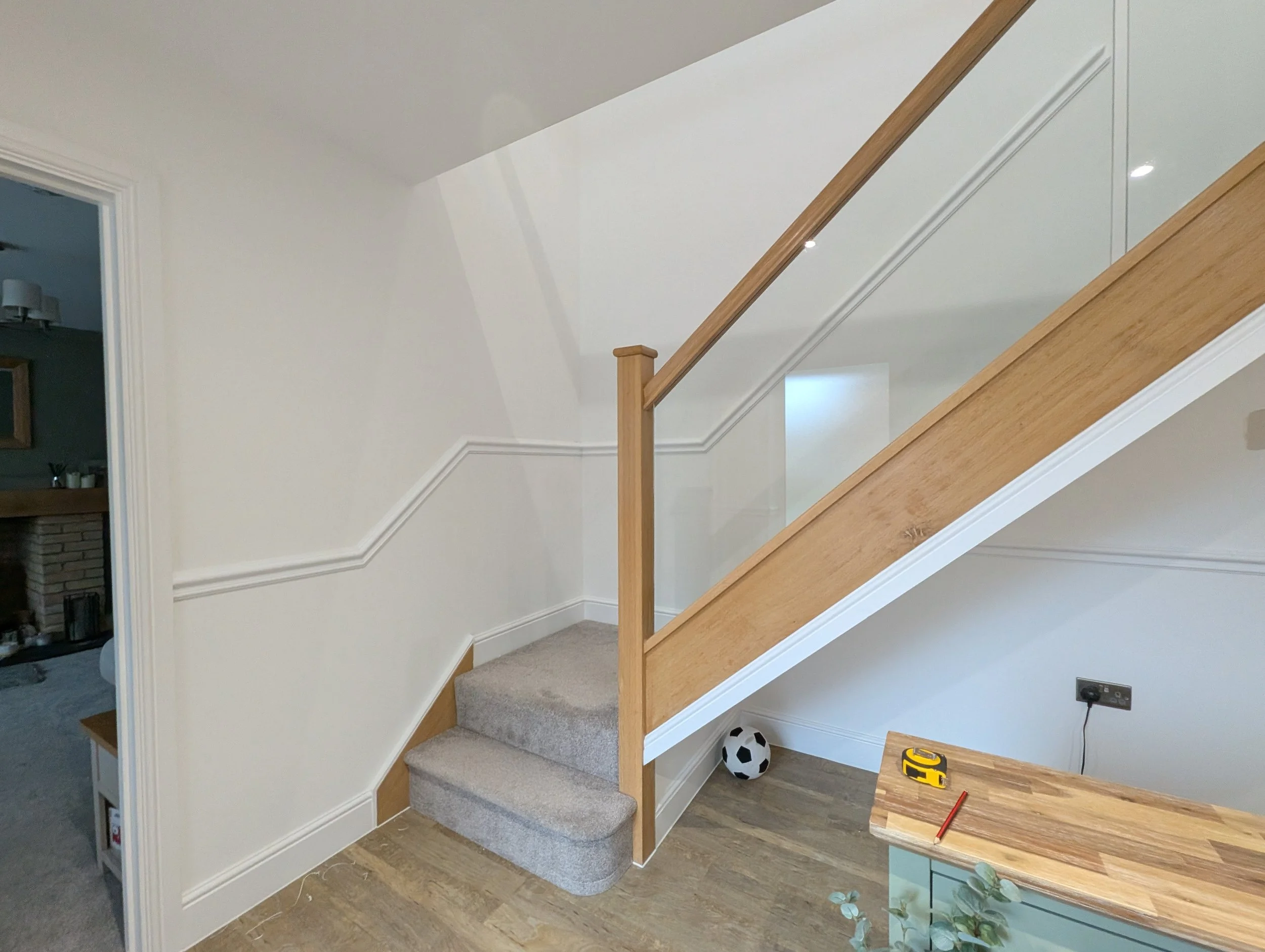 Interior view of a staircase with carpeted steps, wooden railing, and a soccer ball on the floor beneath the stairs. Part of a table with a tape measure and pencil is visible in the lower right corner.