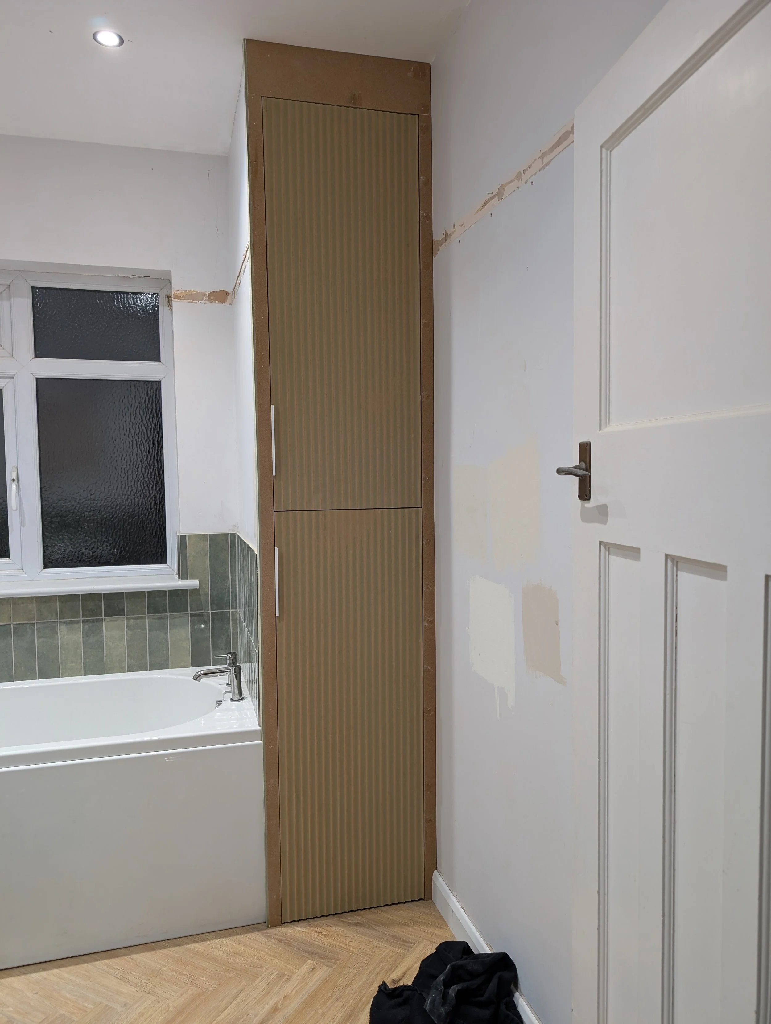 Partially tiled bathroom with a white bathtub, green tiles, and a window, and a tall, ribbed, golden-colored door or panel next to a white door on a wall with visible patches of unpainted wall and a pile of black clothing on the wooden floor.