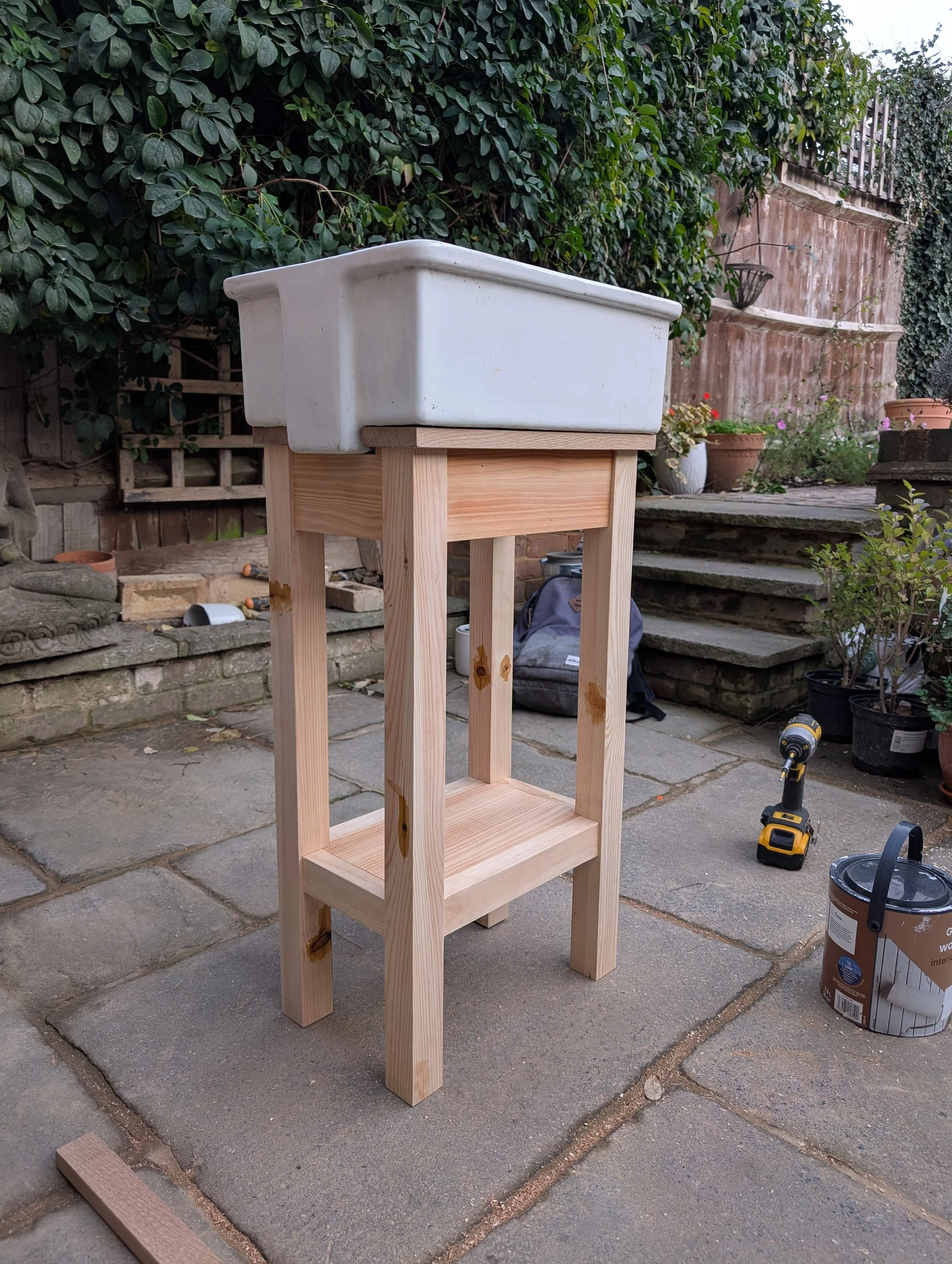 A white rectangular planter on a wooden stand in a backyard with plants, potted flowers, steps, and gardening tools.