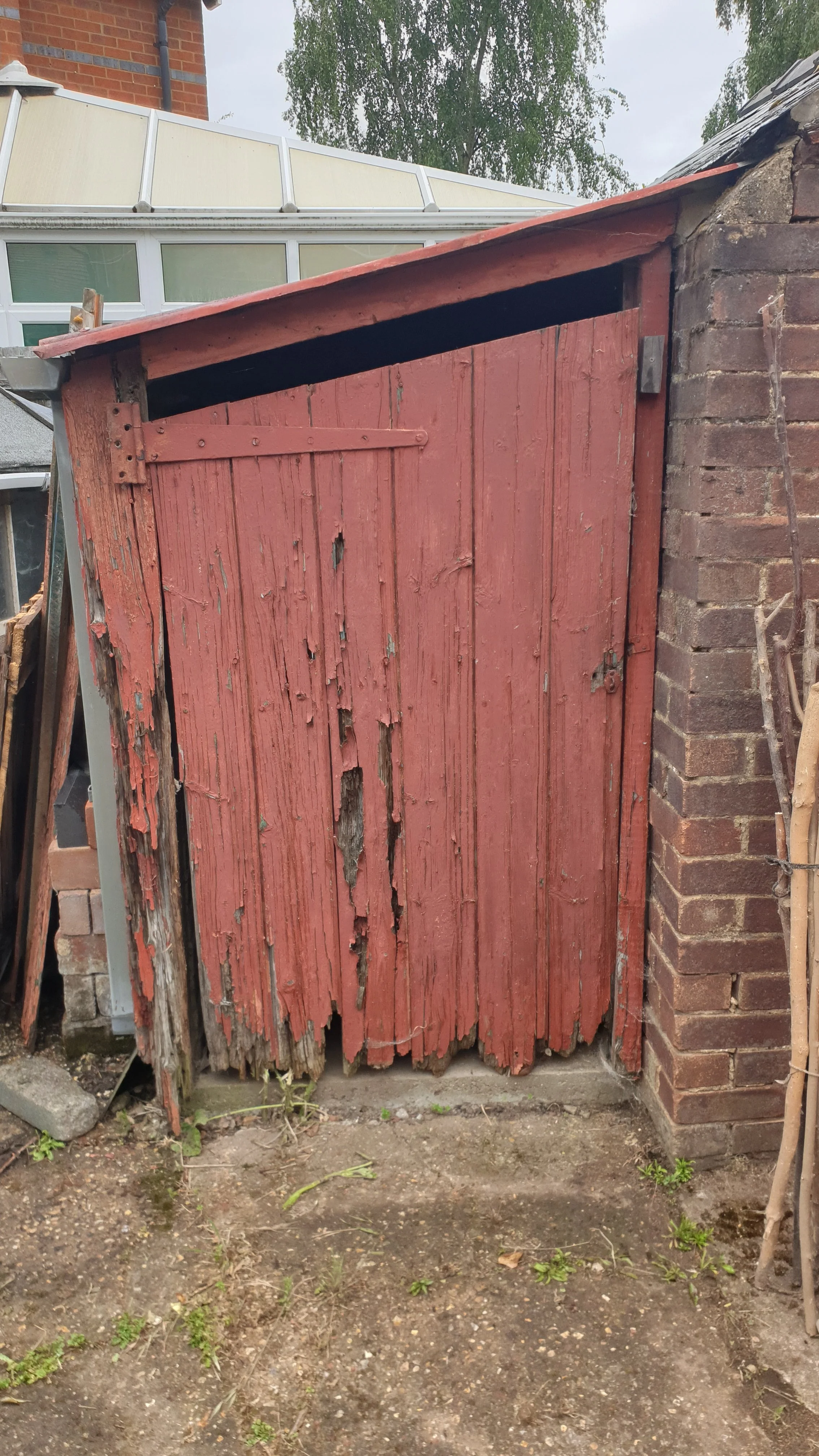 An old, weathered red wooden shed door with peeling paint and rotting wood, situated next to a brick wall, outdoors on a dirt ground.