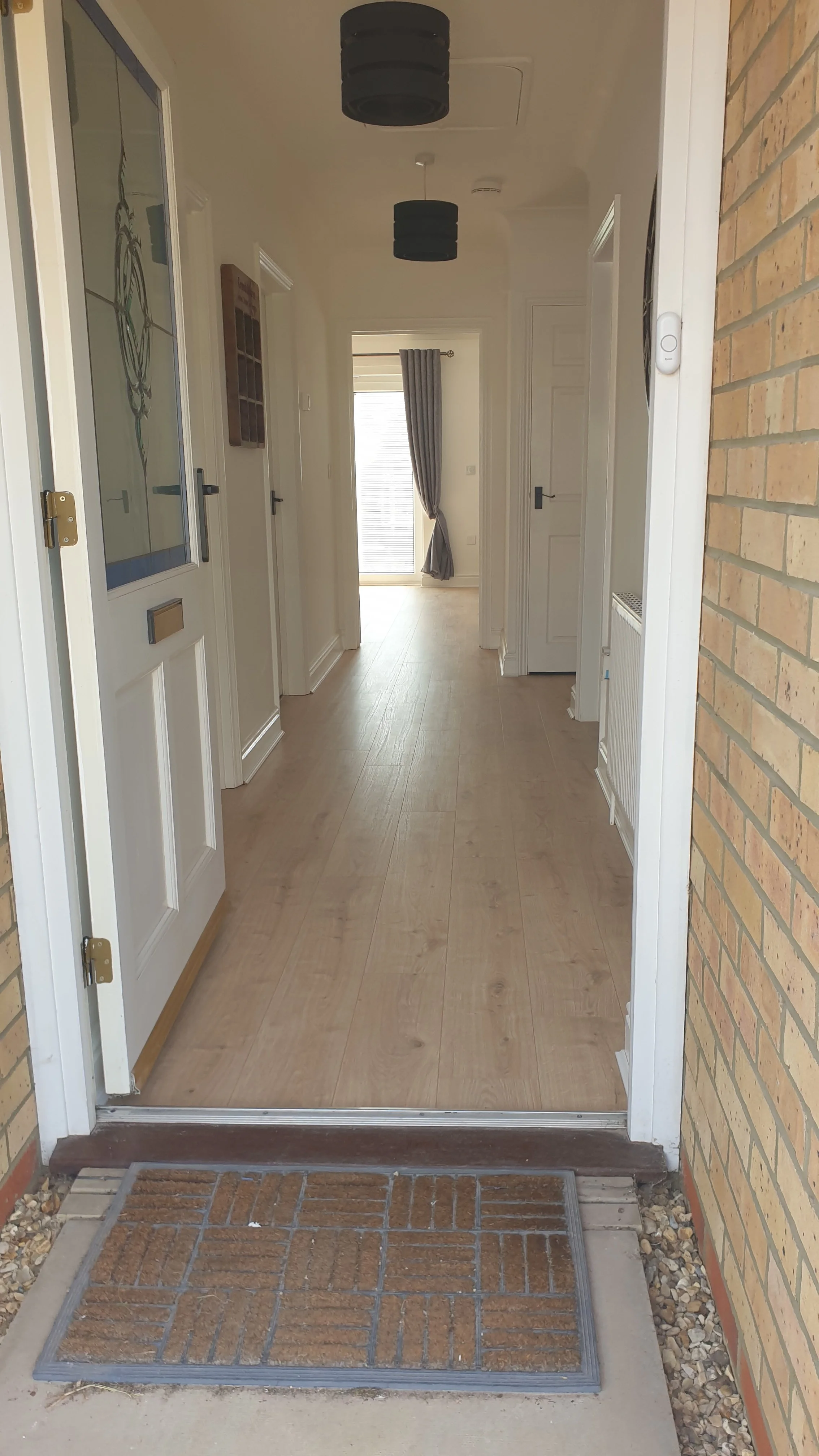 View from the entrance door into a hallway with wooden flooring, white walls, black ceiling lights, and a window with curtains at the end.
