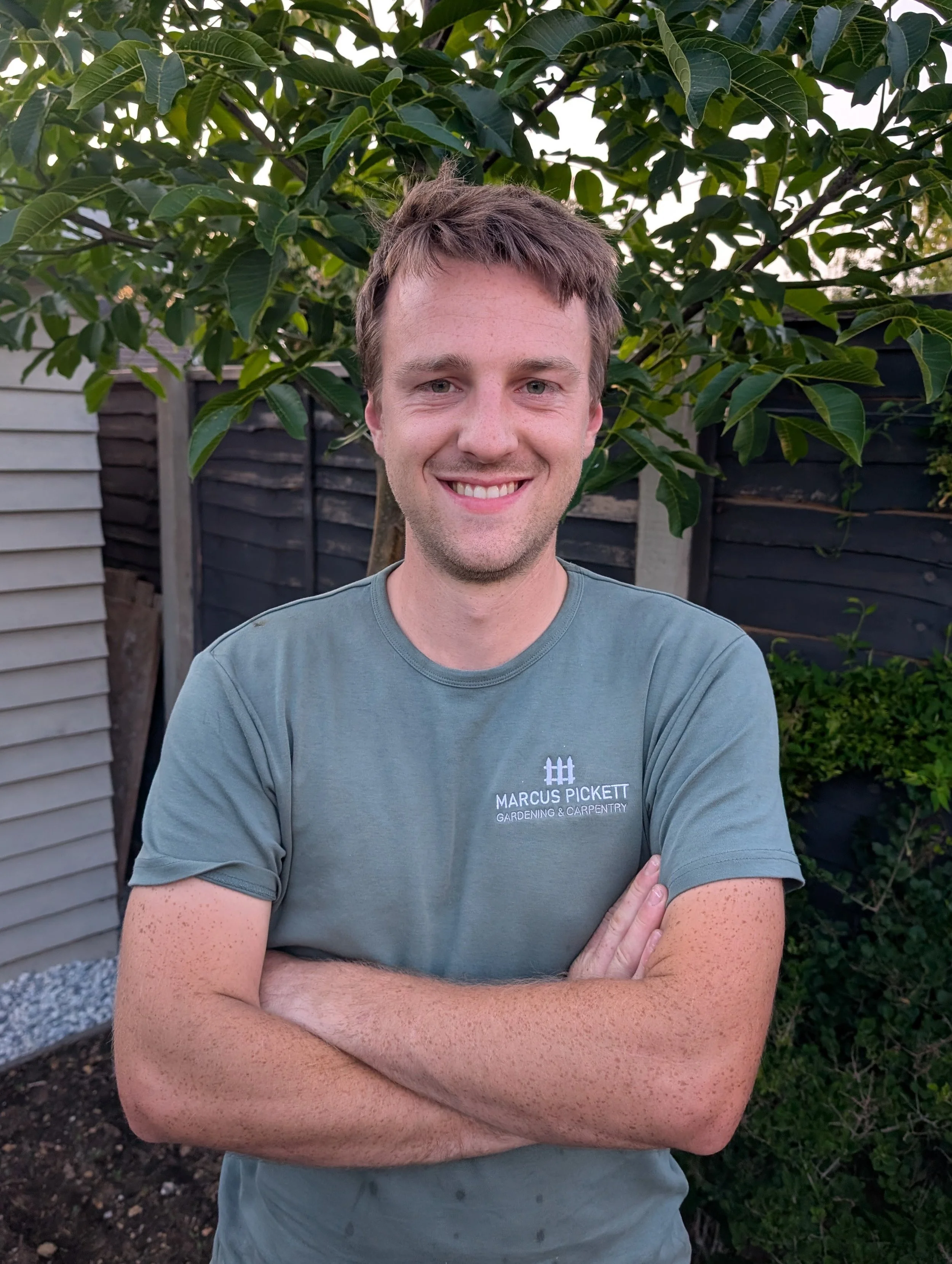 A young man with light brown hair, wearing a grey T-shirt with the text 'Marcus Pickett Gardening & Carpentry,' stands with arms crossed in front of a leafy green tree and a wooden fence in a backyard.