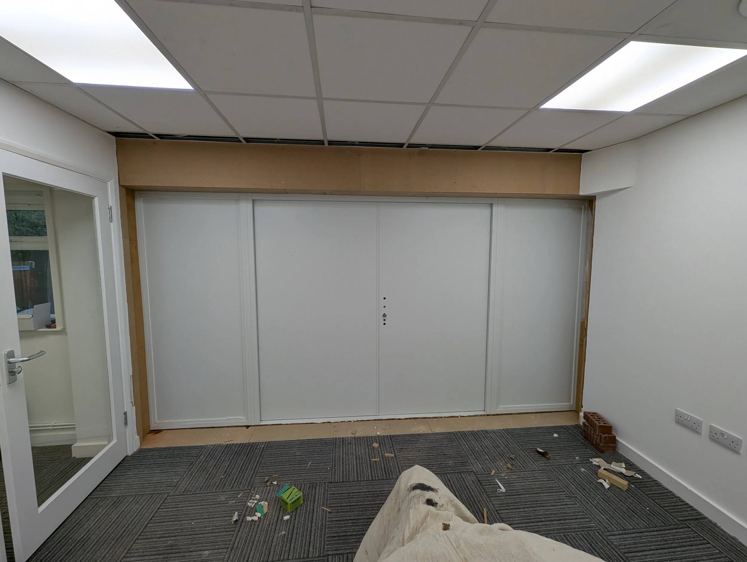 Interior room with white walls, gray carpet, ceiling lights, partially installed wooden frame around a large white sliding door, and cluttered floor with construction debris.