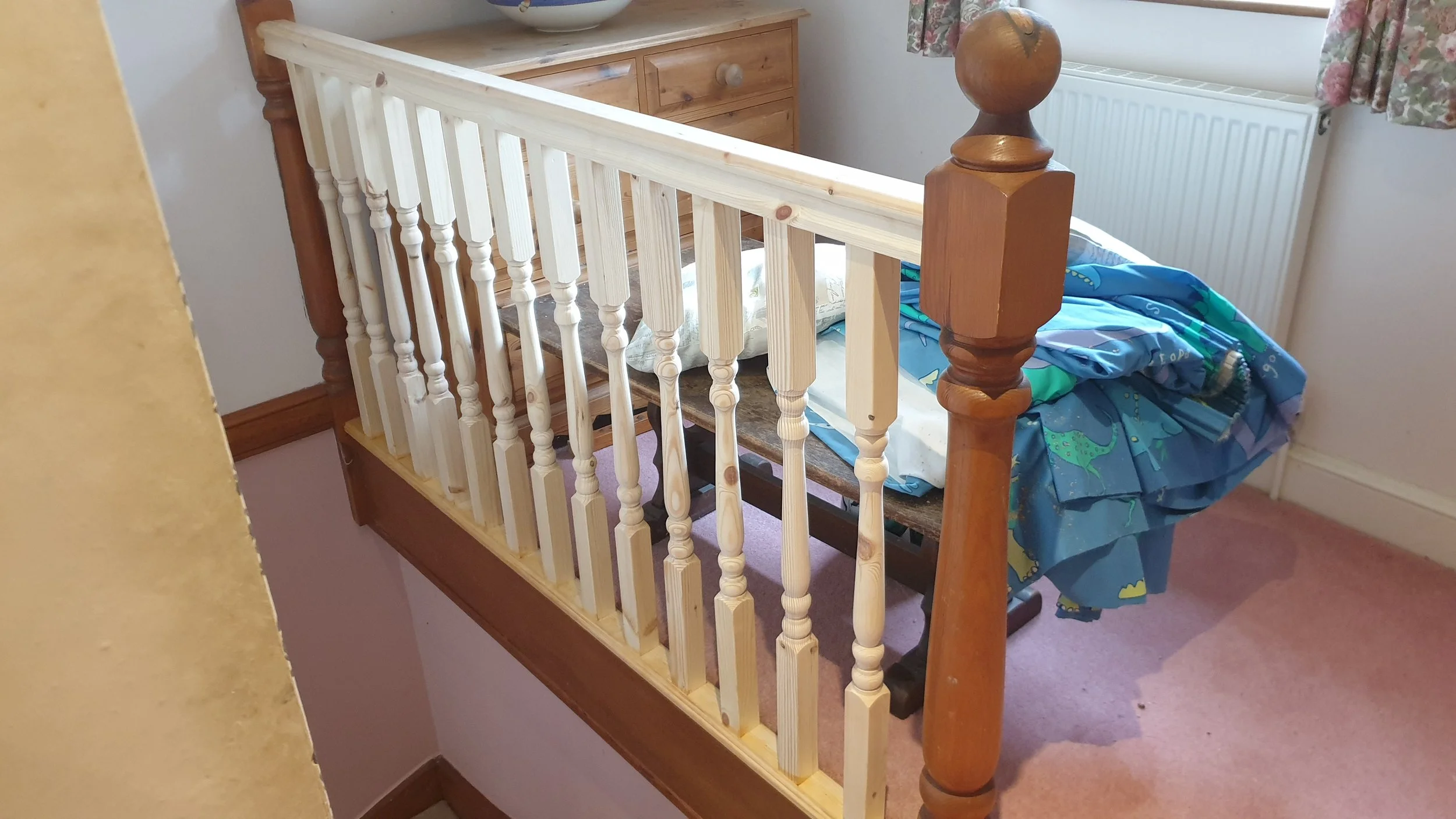 A wooden bed with a colorful bedspread and a pillow on top, situated next to a window with floral curtains, in a room with pink carpet and a white radiator.